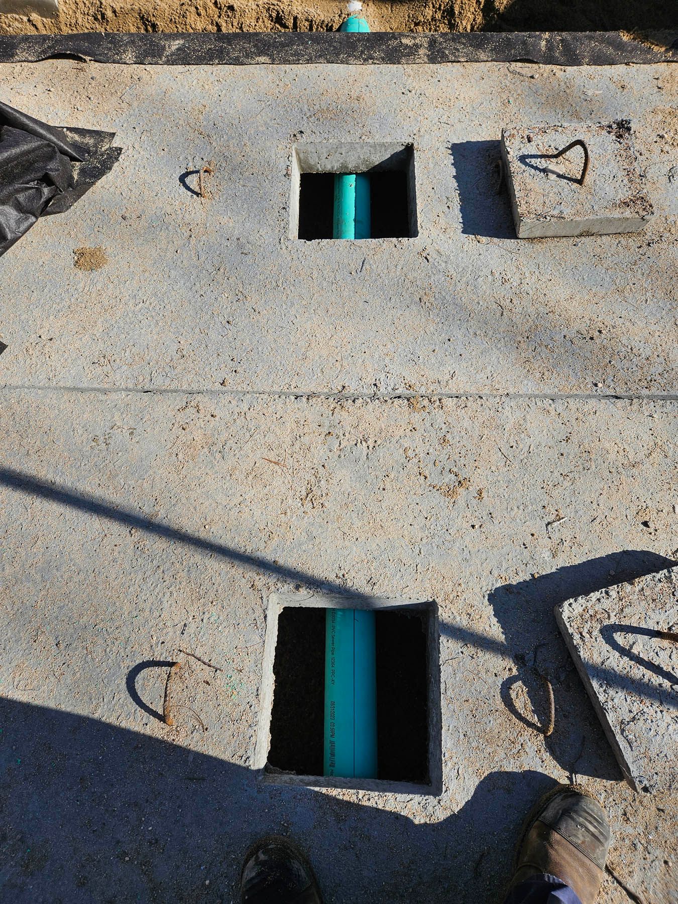 Concrete septic tank lid with two square openings for pipes. Green pipes visible.