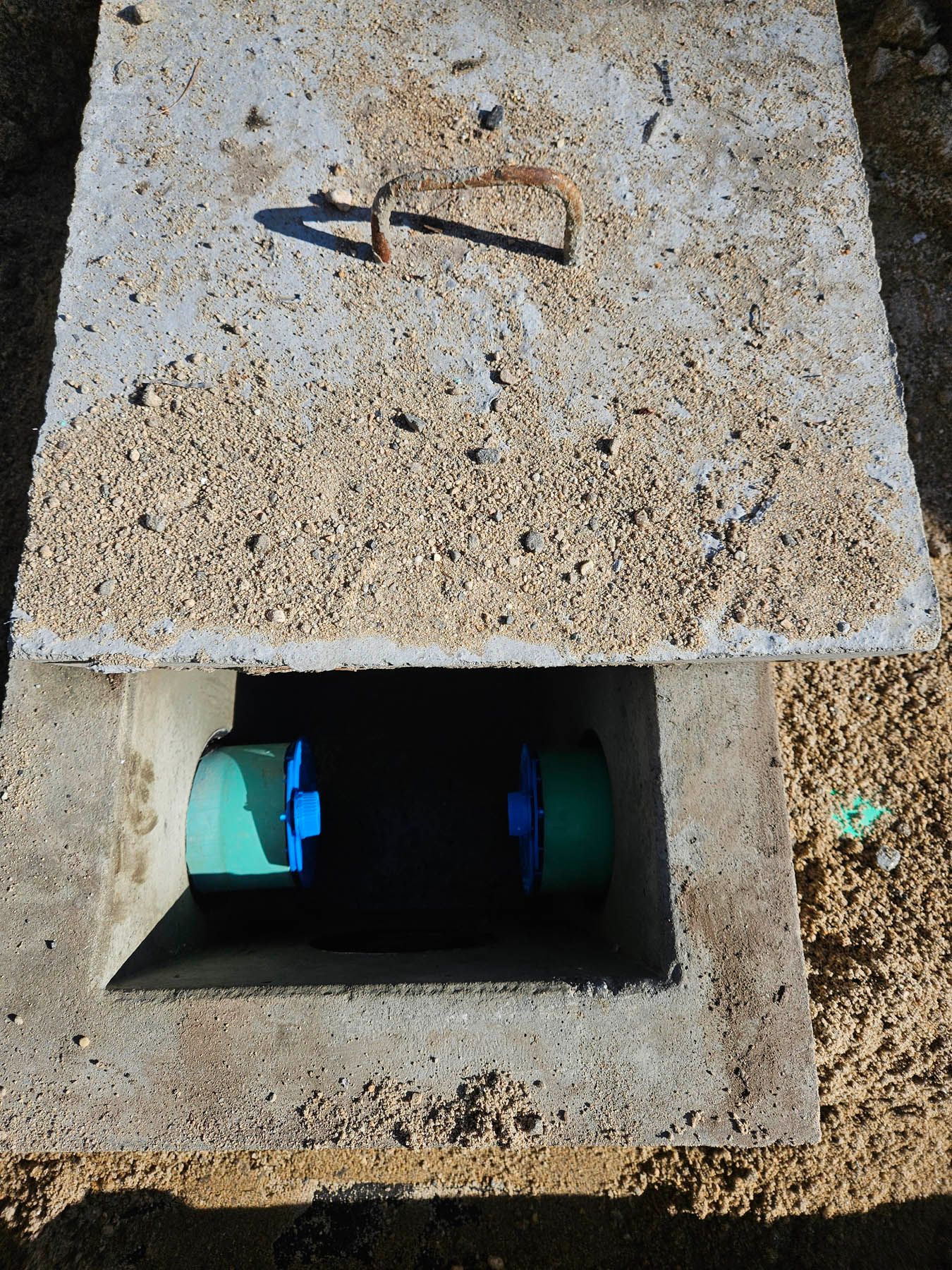 Concrete utility access point with cover removed, revealing green pipe couplings.