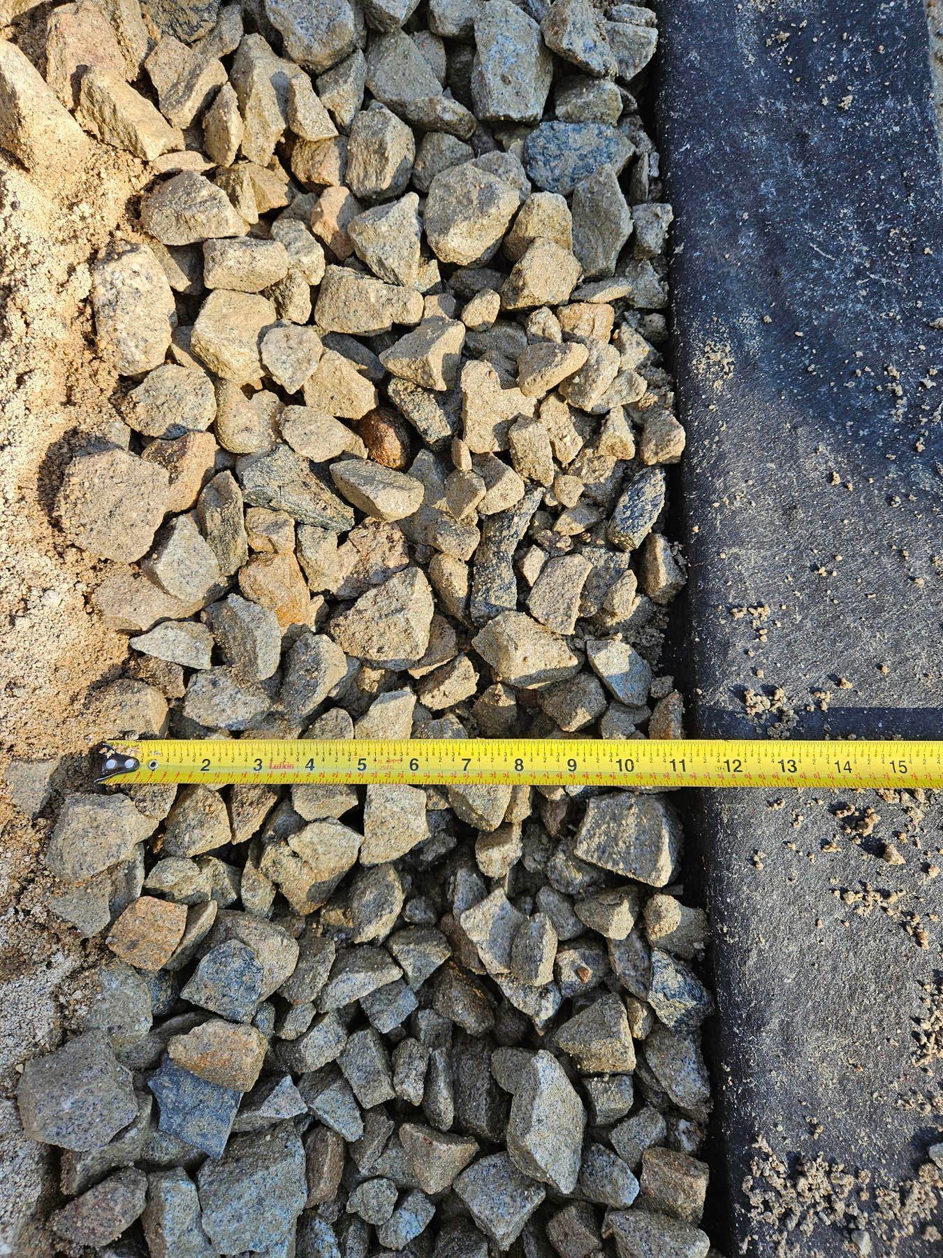 Gravel and sand next to black landscape fabric, with a yellow tape measure showing width.