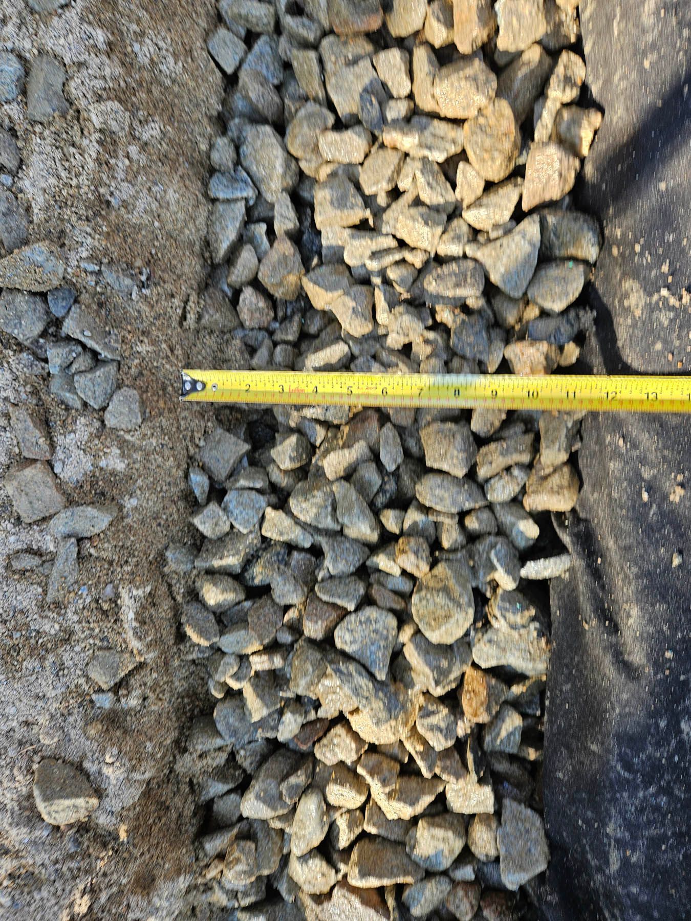 A tape measure spanning a rock-filled trench, likely for drainage, next to a sand-like surface.