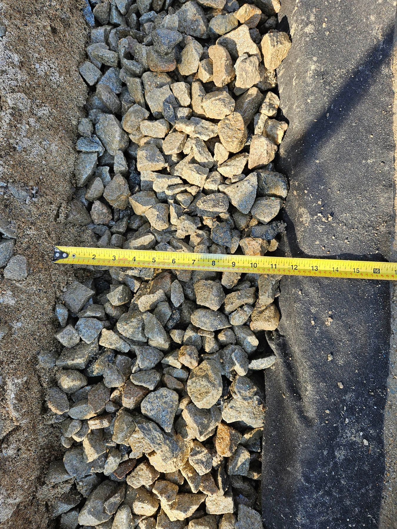 Gravel-filled trench with a measuring tape. Ground is tan and black.