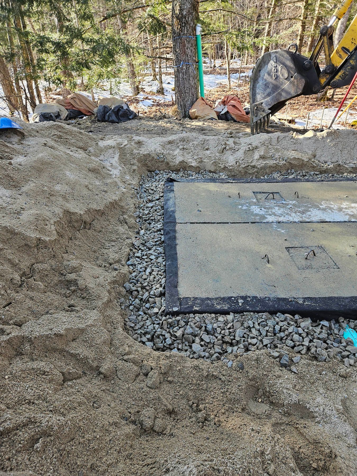 Excavation around a concrete slab with gravel border; backhoe in the background; snowy woods.