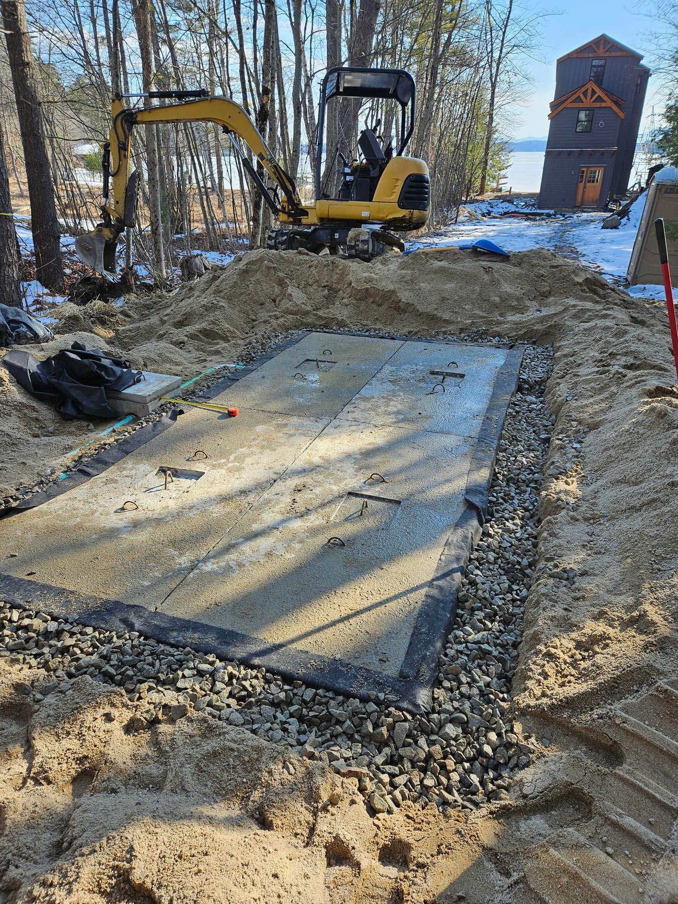 Mini excavator working on a rectangular concrete foundation in a sandy, wooded area.