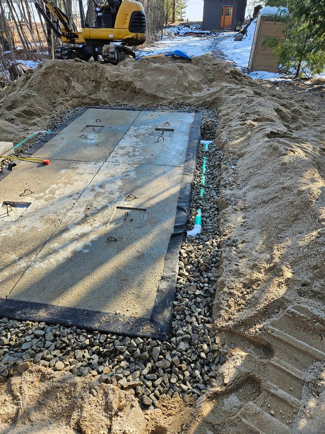 Excavation site with a concrete septic tank, gravel, and piping in a yard with snow.