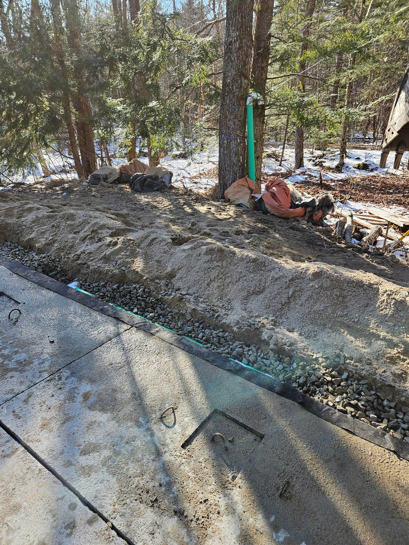 Construction site with soil, gravel, and tools next to a paved area. Trees in the background.