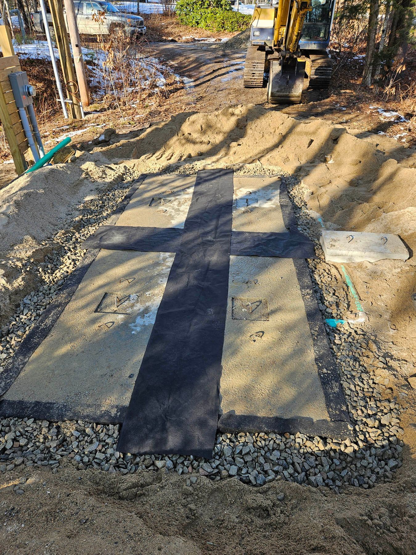 Concrete pads in a cross shape, connected by black material, within an excavated area, with a small excavator in the background.