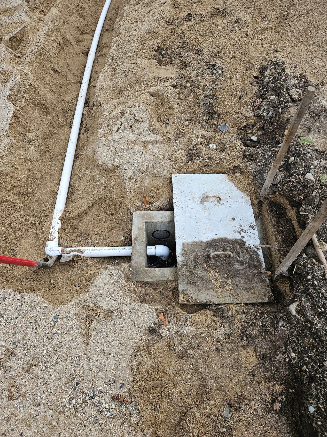 A partially buried septic tank with a concrete cover, connected to white PVC pipes in a dirt trench.