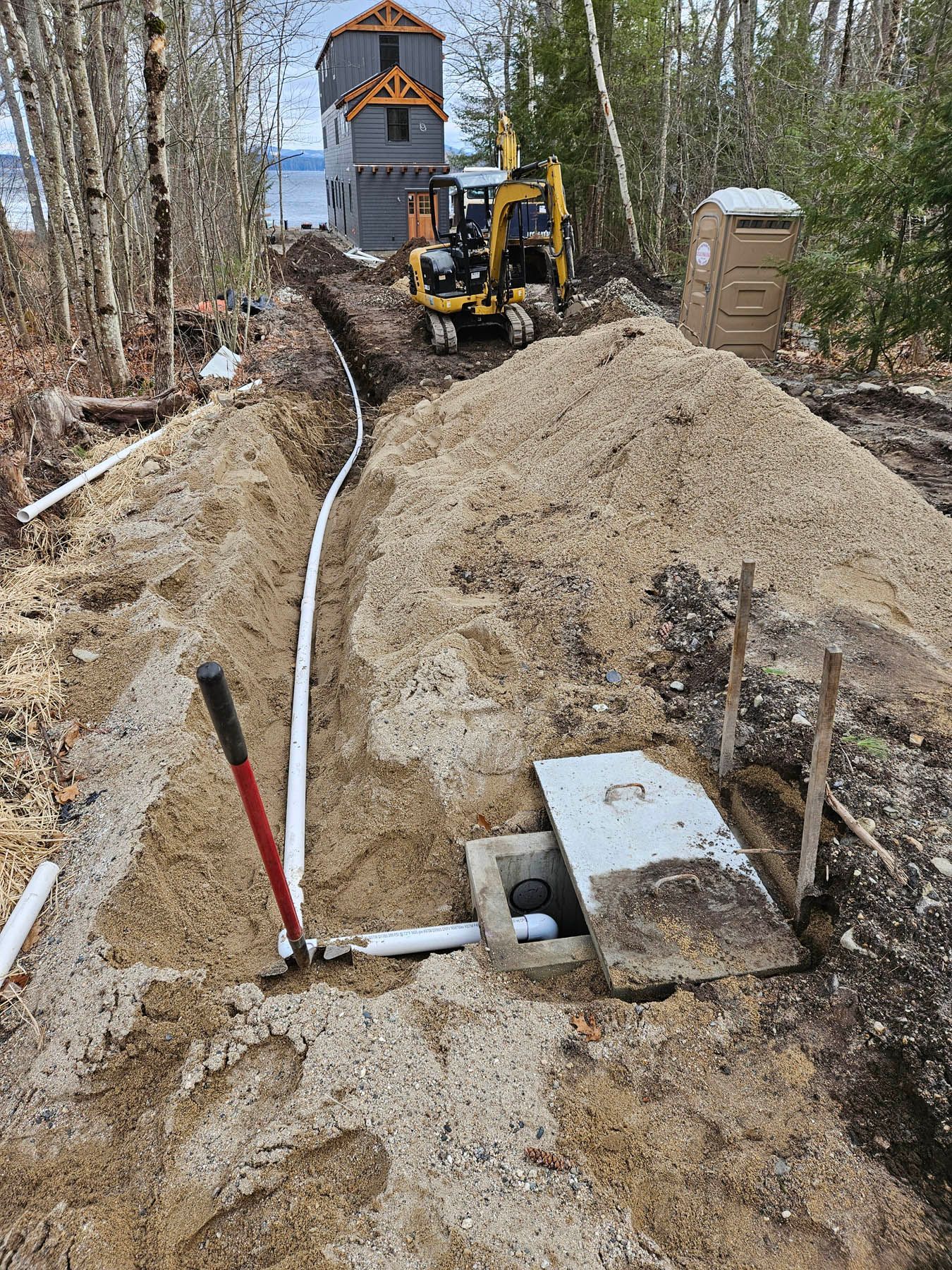 Construction site: Excavator digging trench for pipe, near a building and a portable toilet.