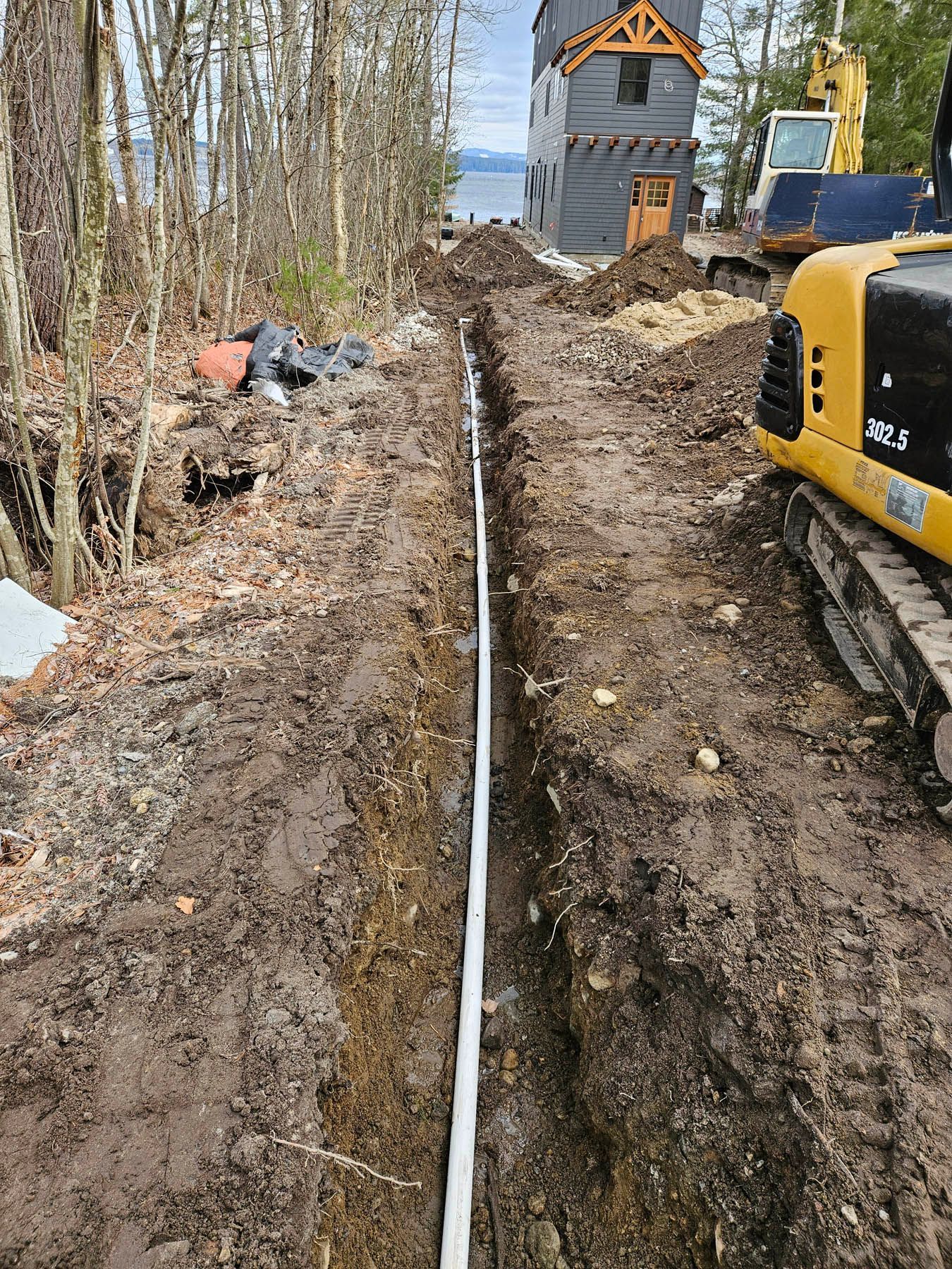 A trench with white pipe, next to trees and a building under construction.