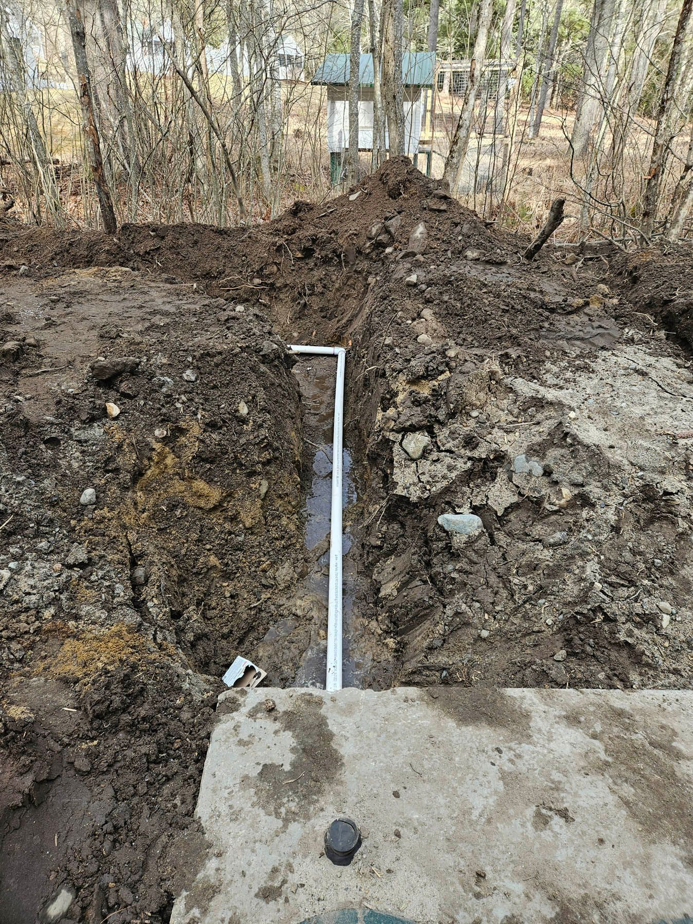 Trench dug in brown soil, with a white pipe in it. Concrete slab in foreground; trees in the background.
