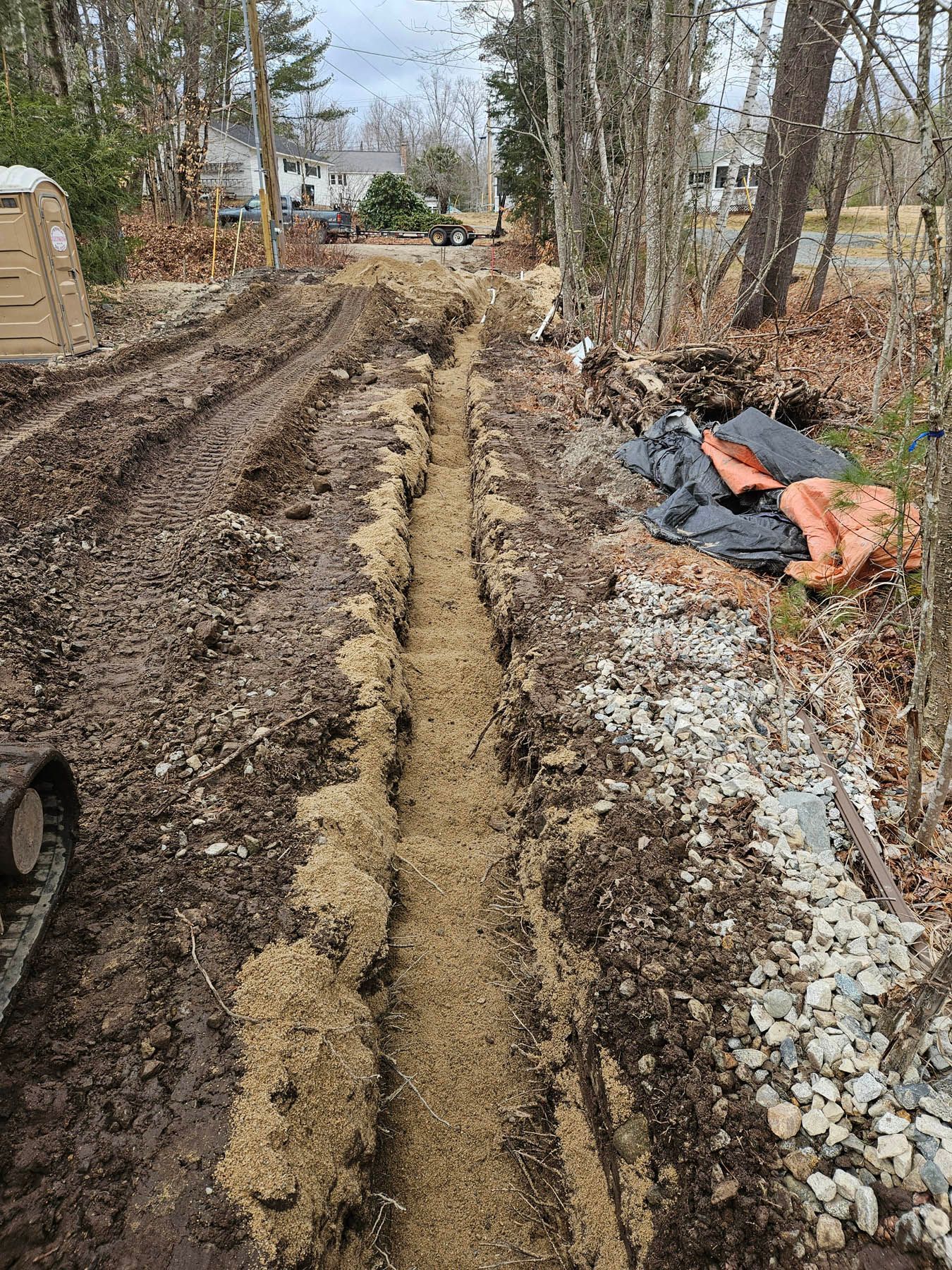 Trench dug in muddy ground, with sand lining the bottom; trees and buildings in the background.