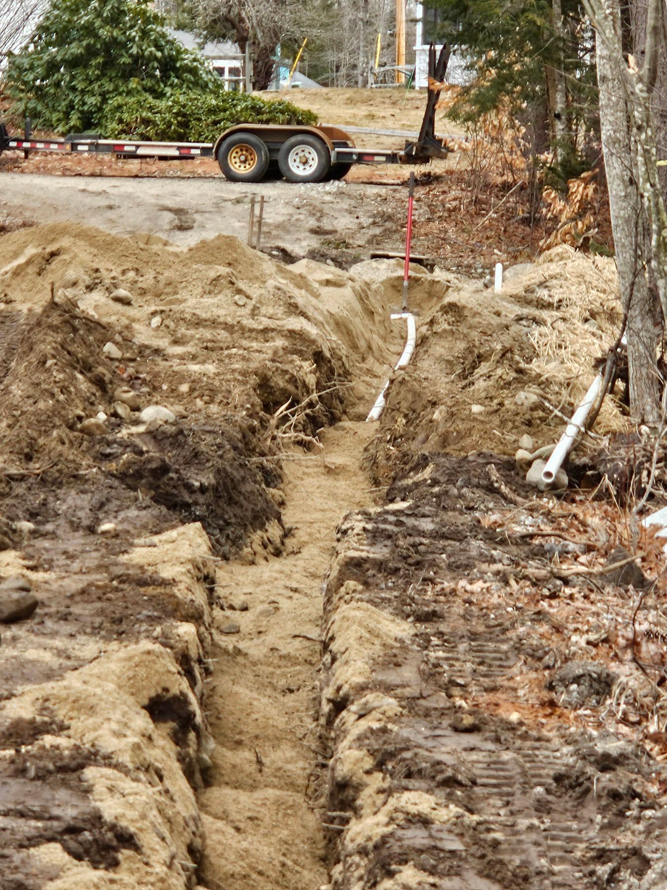 A long, narrow trench dug in sandy soil, with white pipes running through it, likely for utility installation.