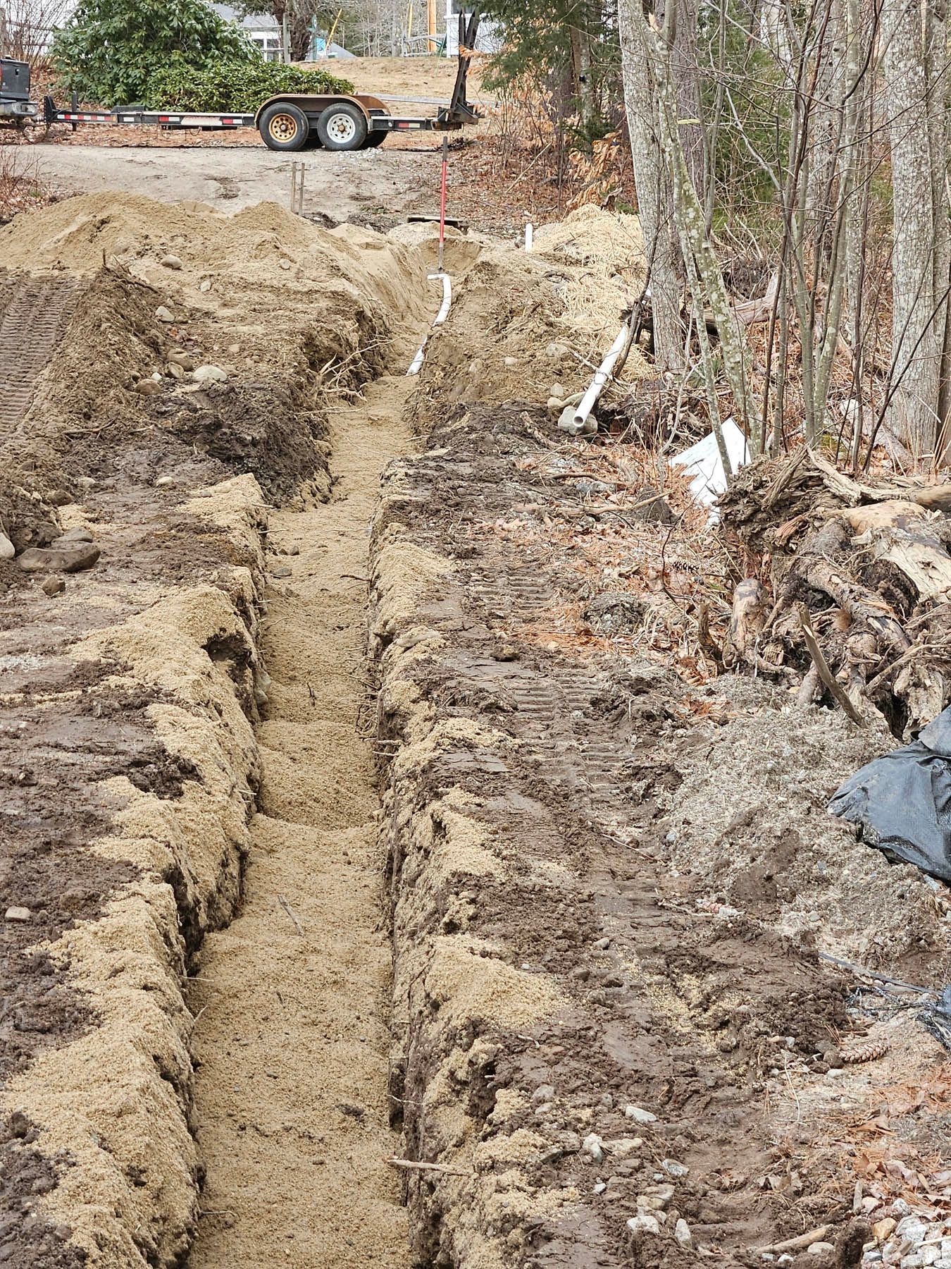 A trench dug in muddy ground with white pipes visible, likely for utilities, near trees and a lawn.