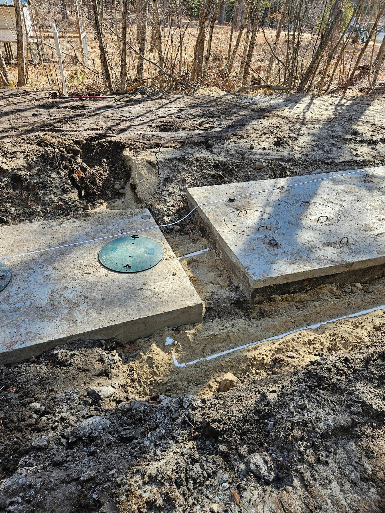 Two rectangular concrete septic tank lids sit in a dirt excavation, with connecting pipes visible.