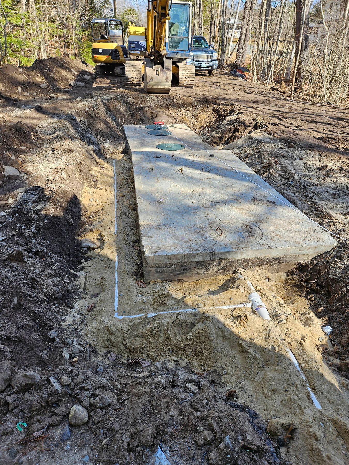 Excavator installing a concrete septic tank in a construction site; dirt, pipes, and machinery visible.