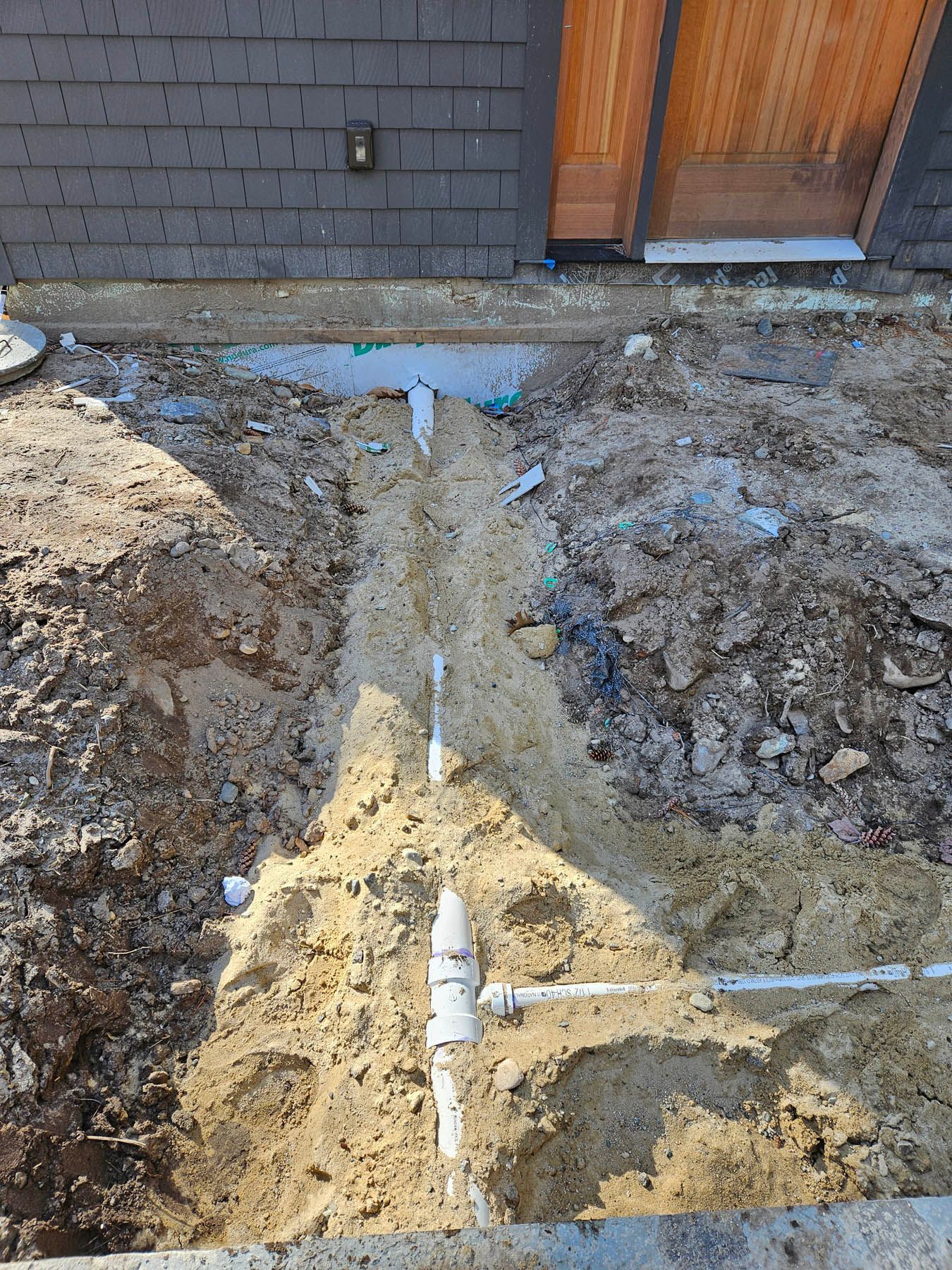 Trench with white PVC pipes installed near a building's foundation; a wooden door is visible.