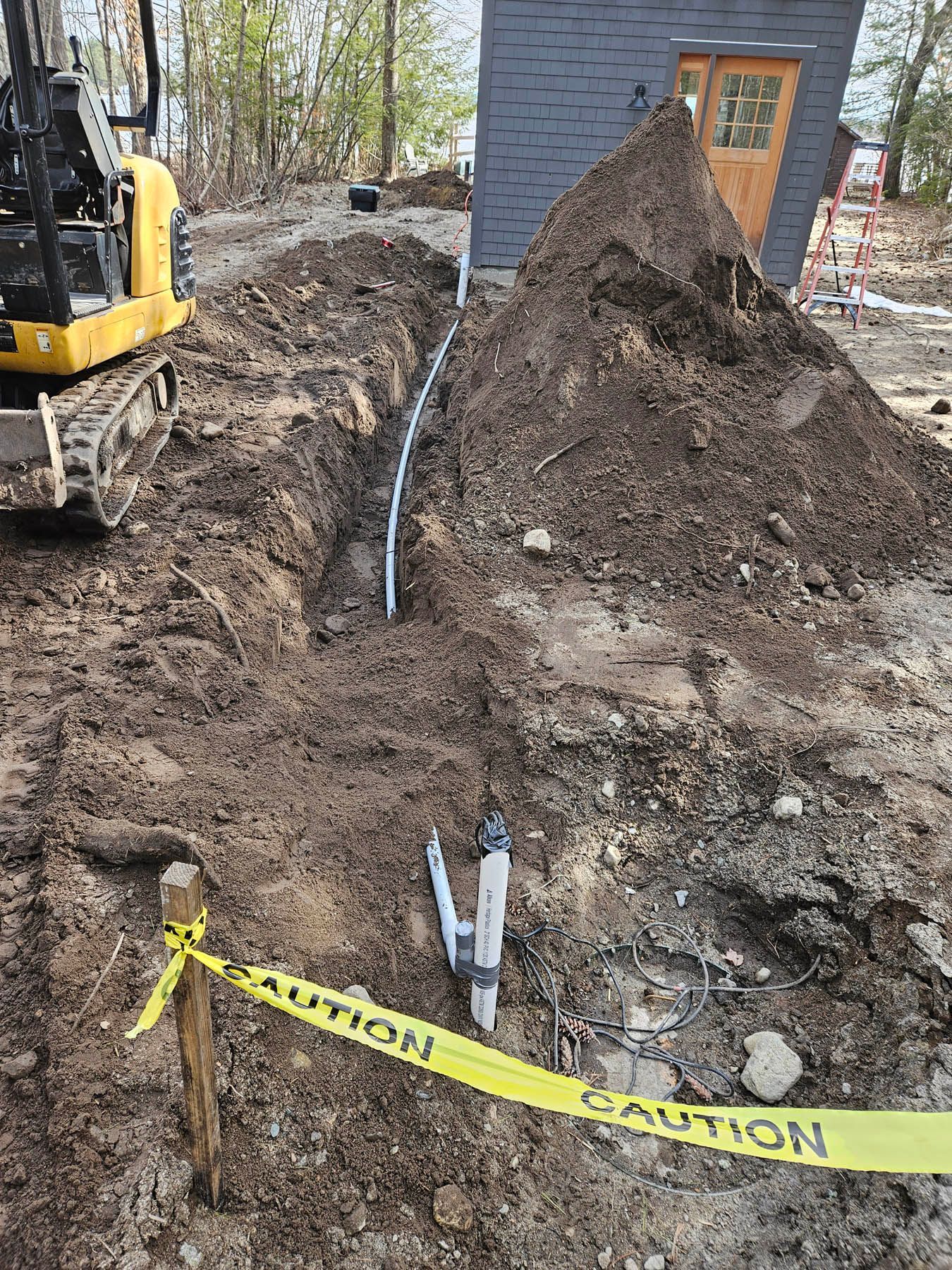 Trench with buried pipe near a building under construction. Yellow caution tape and an excavator are present.