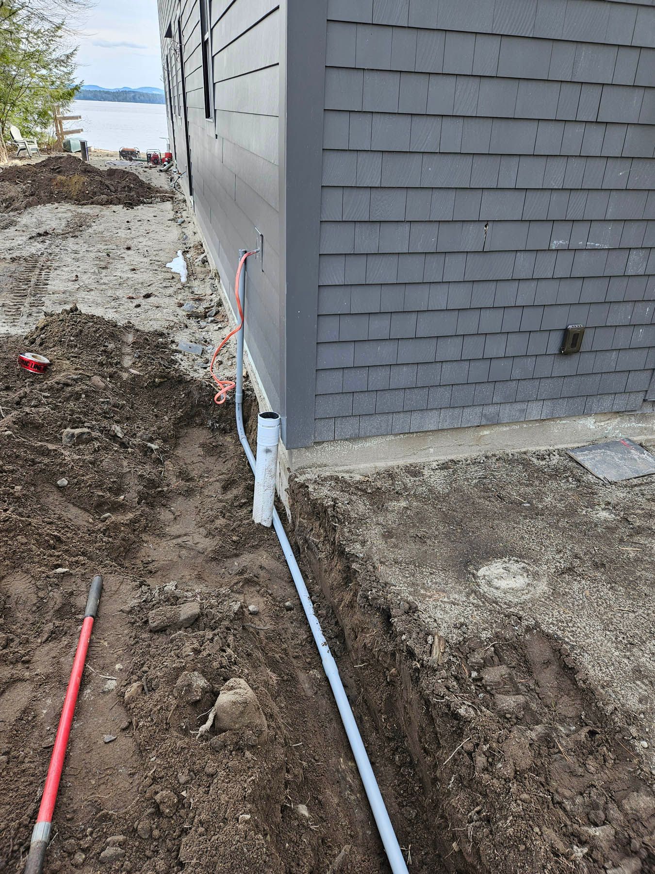 Trench dug near a gray building with exposed pipes, red shovel, and dirt.