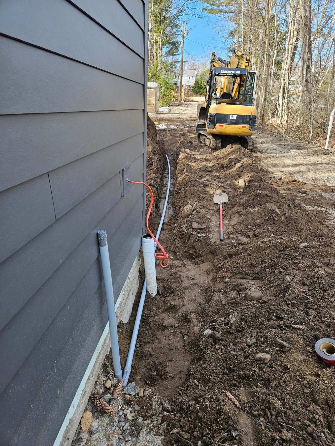 An excavator digs a trench near a gray building, with conduit and orange wires visible.