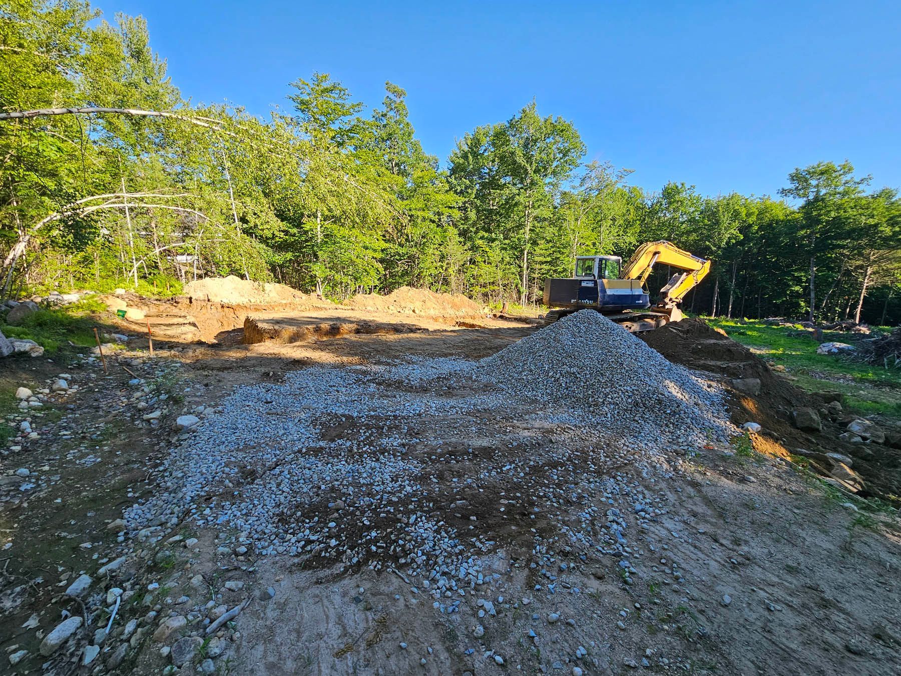 Construction site with a pile of gravel in front of a small excavator, clear sky, trees in background.