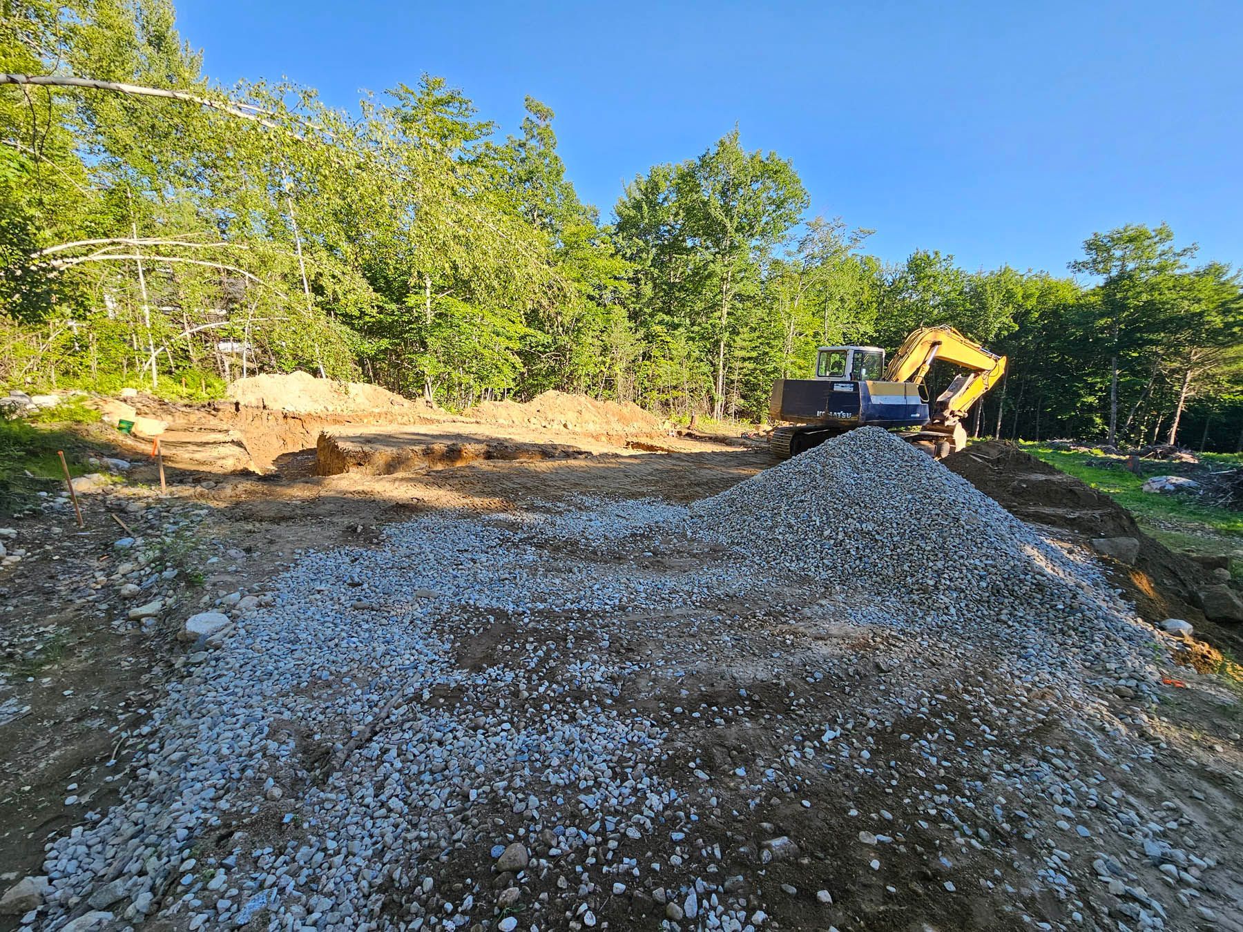 Excavator on a gravel pile, clearing land for construction, with trees in the background under a blue sky.