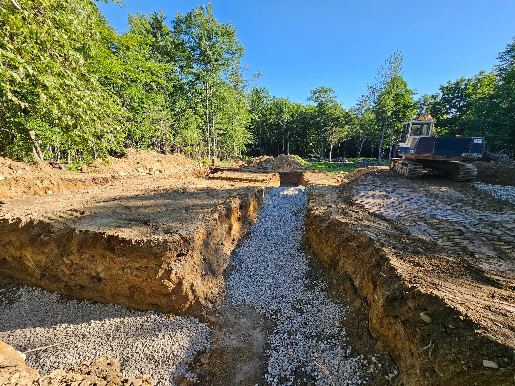Trench dug for foundation with gravel base, excavator in the background; in a wooded area.
