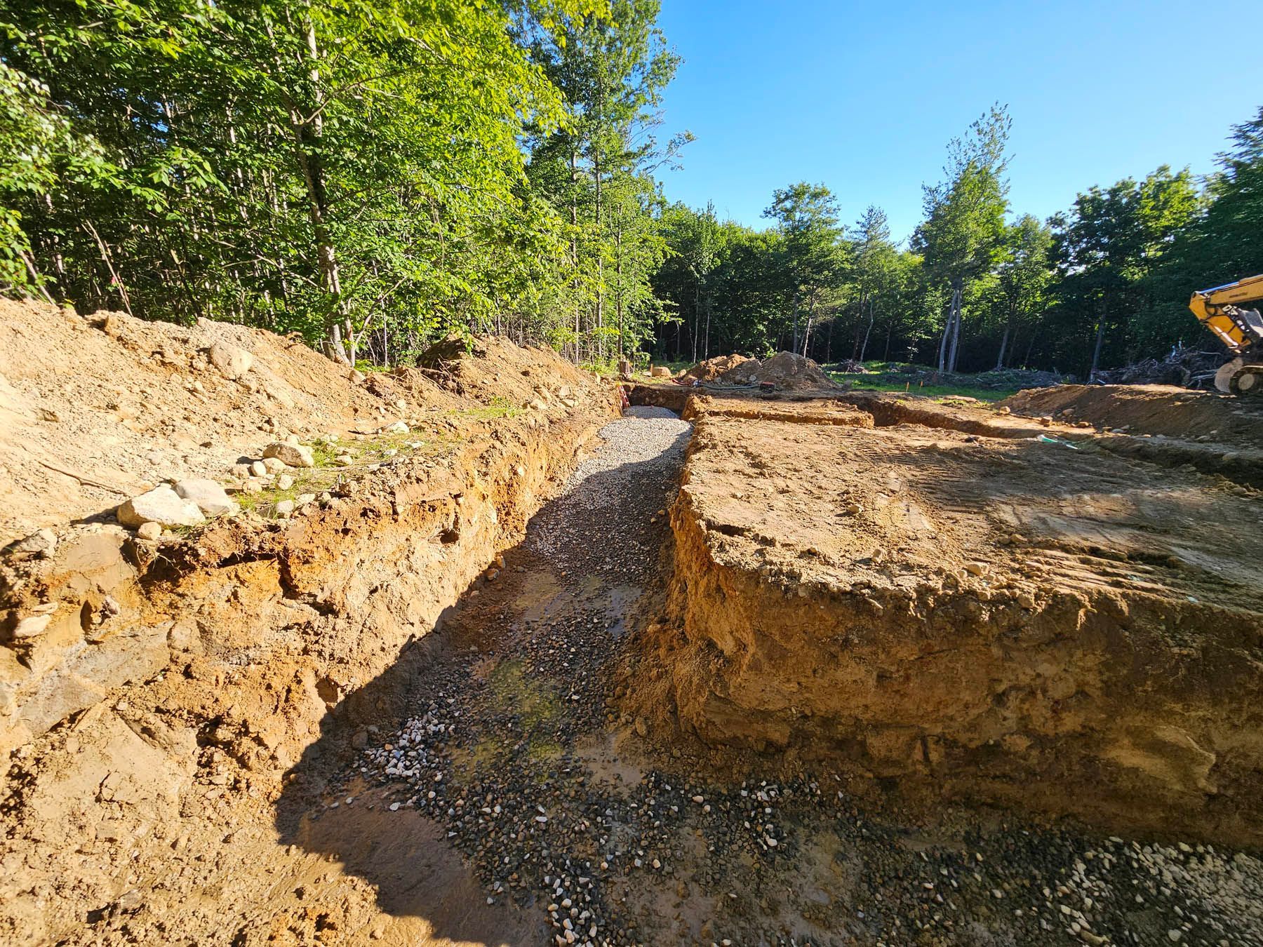 Excavated trench filled with gravel in a wooded area, likely for a foundation.