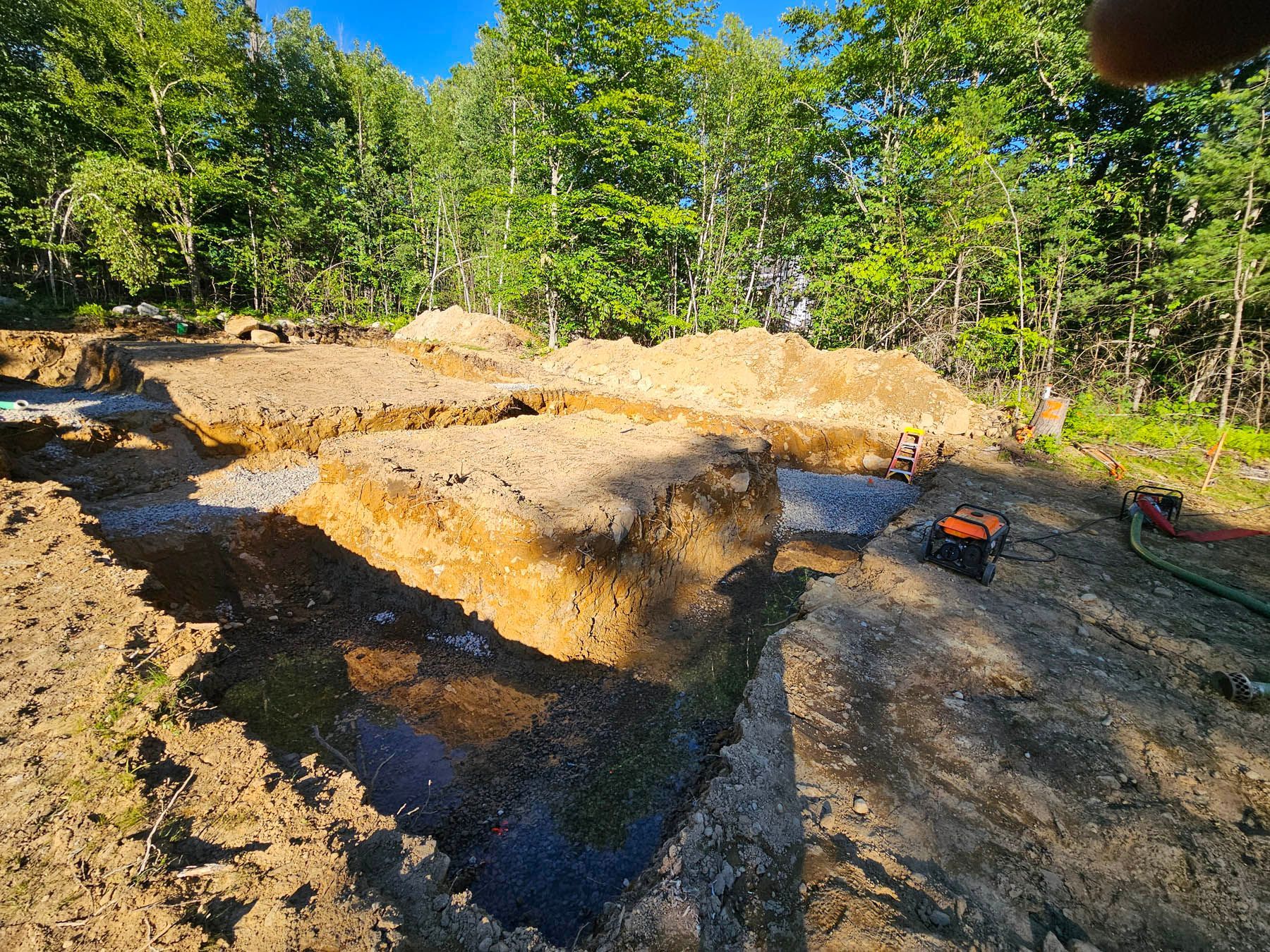 Excavated foundation trenches for a building, with piles of dirt, gravel, and trees in the background under a blue sky.