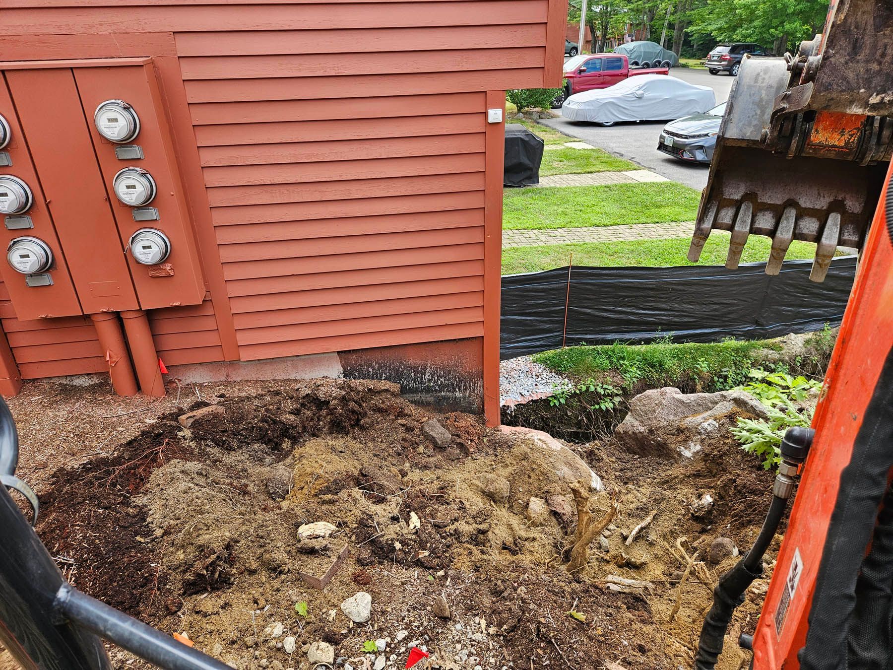 An excavator digging near a red building with electrical meters, brown earth, and a street view.