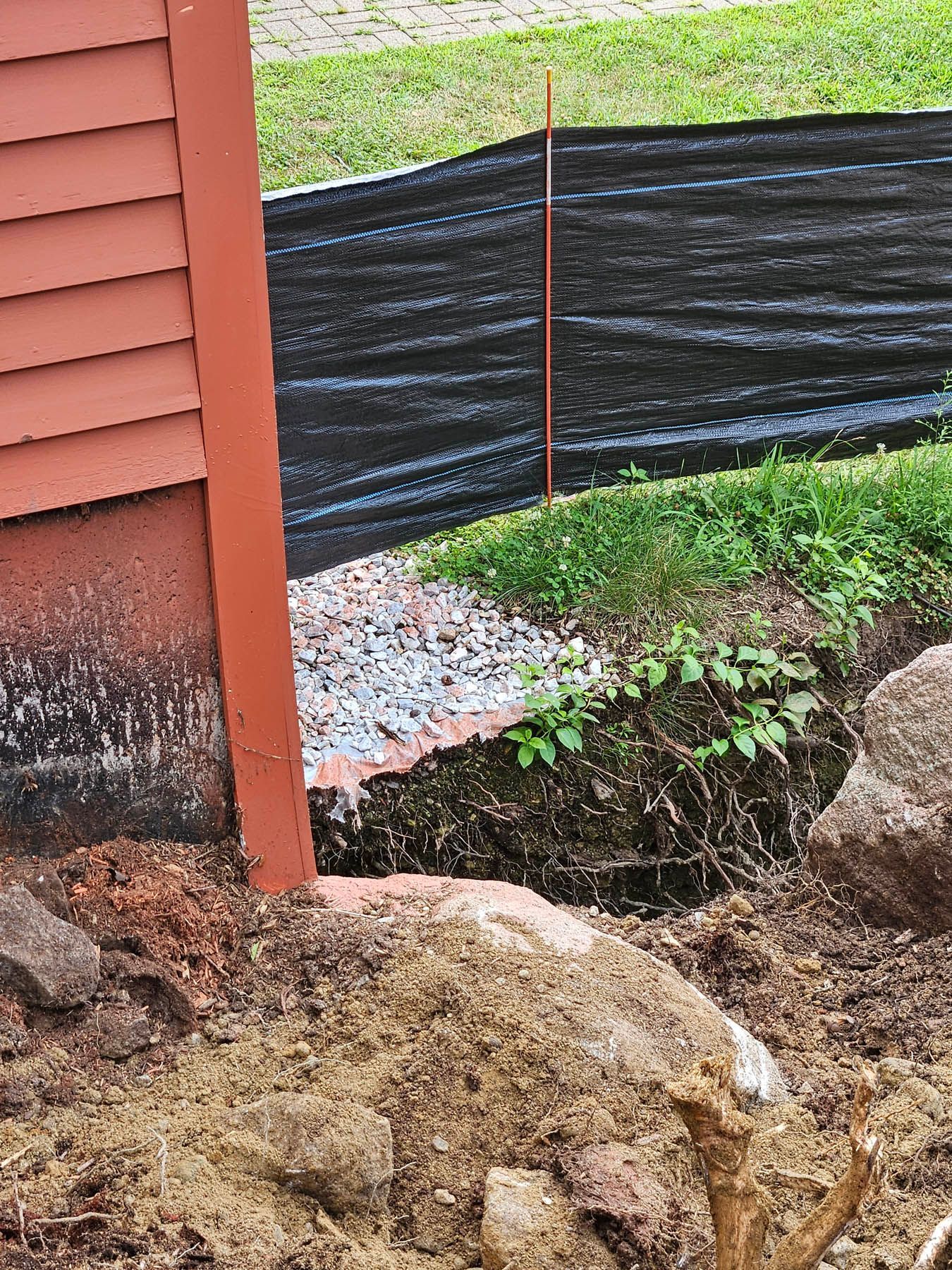 Red building side with exposed foundation, rock barrier, and black fabric.
