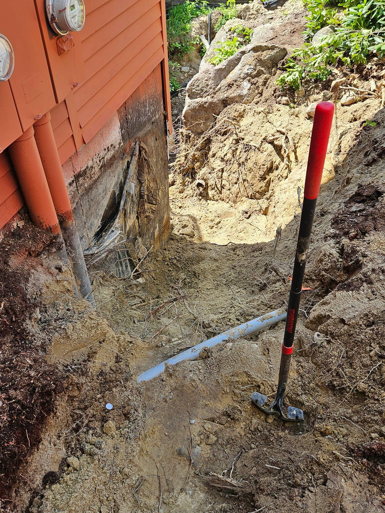 Trench dug next to a building's wall. Shovel leans in the trench; pipes visible, revealing foundation.