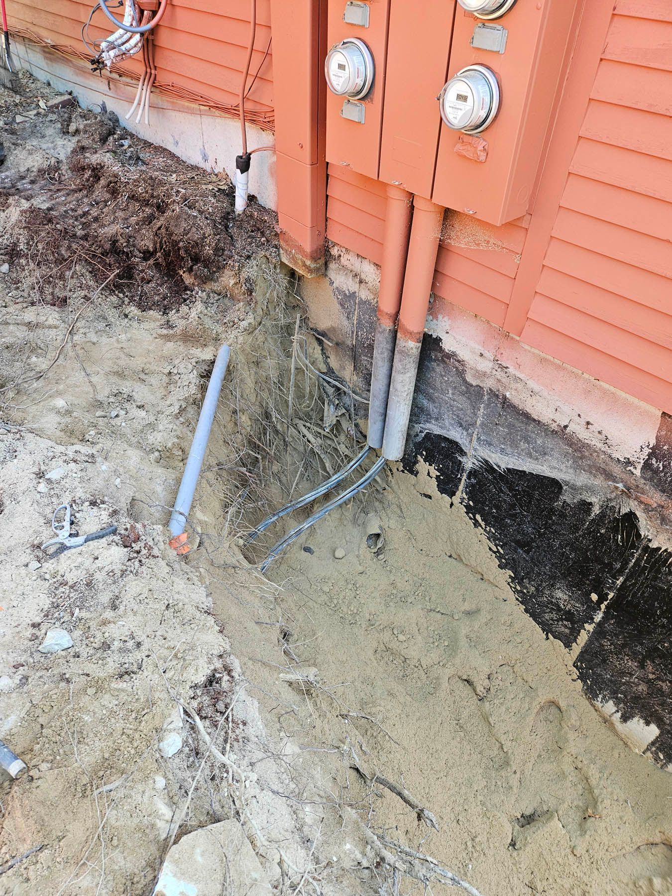 Electrical meters and conduits on a building, with a trench in front.