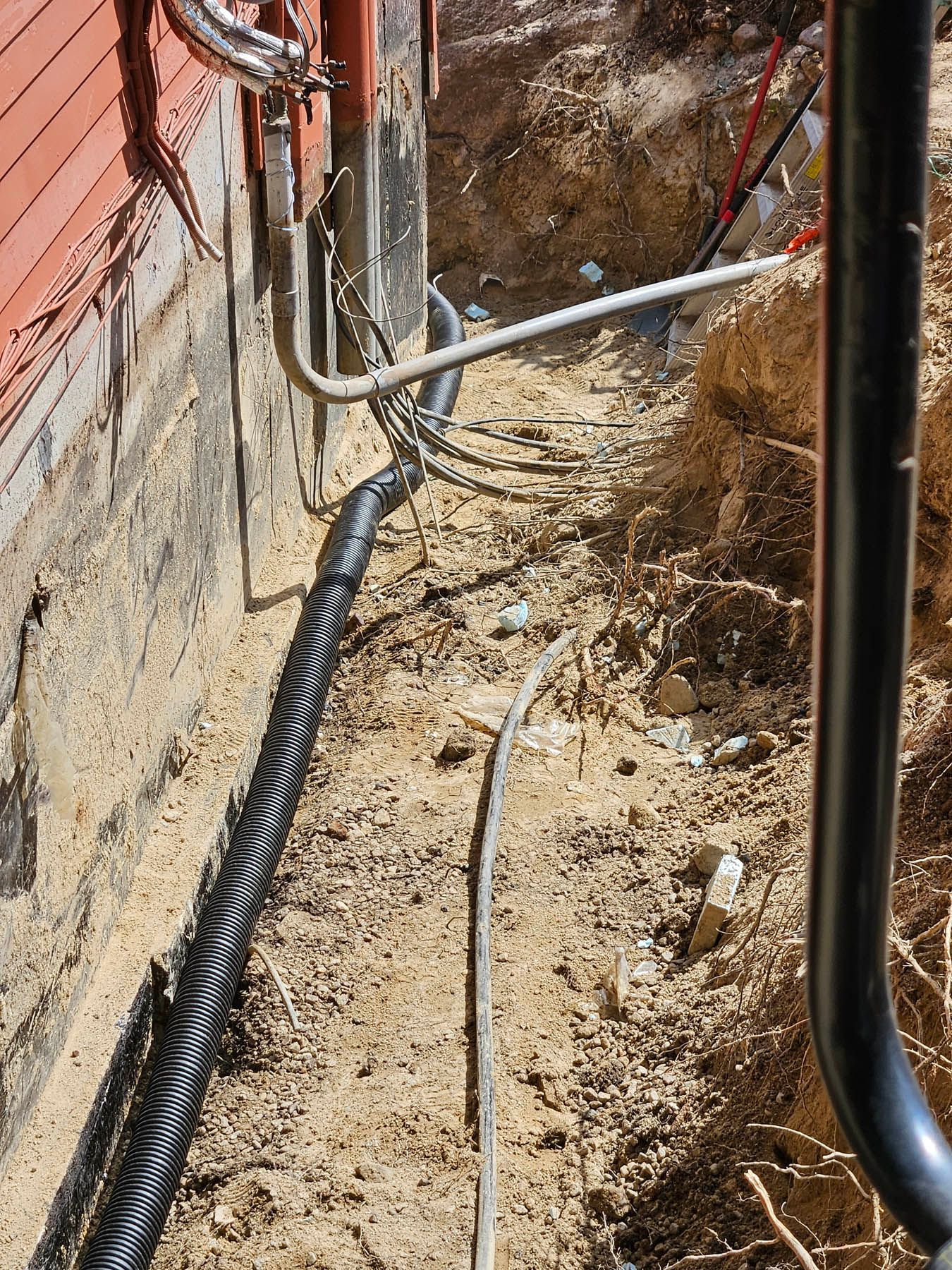 A trench next to a red building with black corrugated drainage pipe and various utility lines.