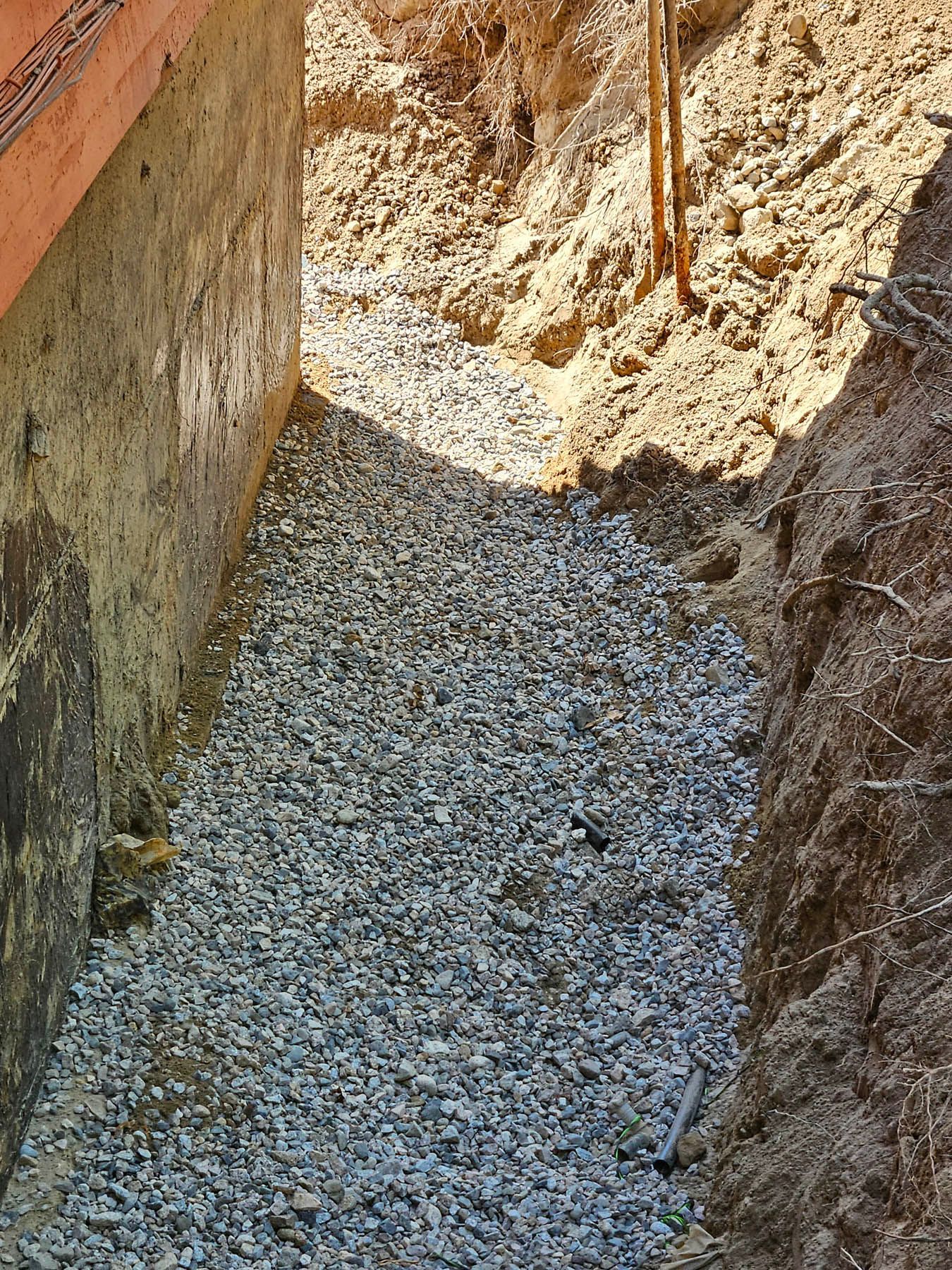 Gravel-filled trench next to a concrete wall and a dirt bank, likely for drainage or foundation work.