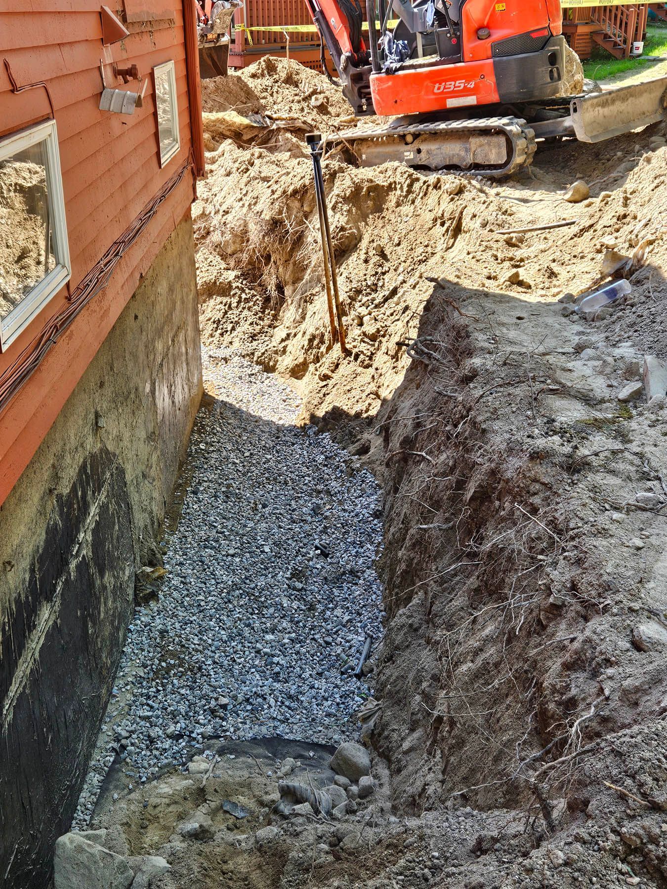 Excavation beside a red building. A trench filled with gravel, machinery and dirt.