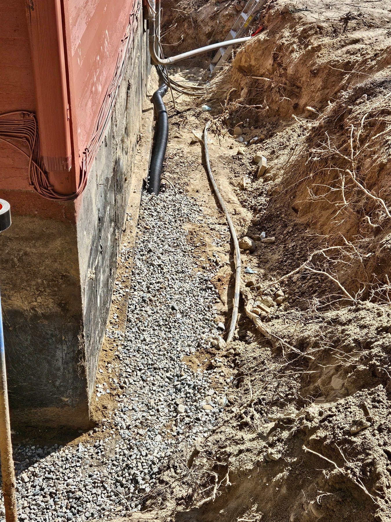 Gravel drainage system against a red building foundation. Black pipe and electrical wires visible in the trench.