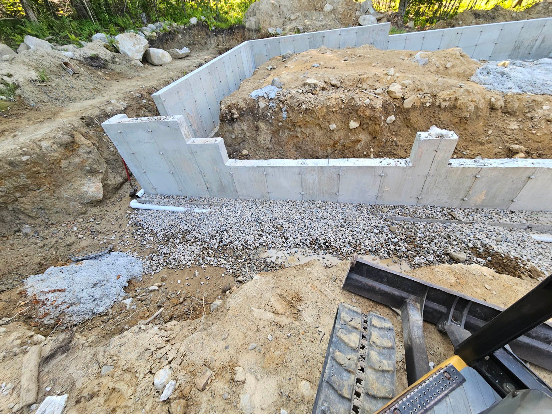 Foundation walls of a building under construction, with gravel and earth visible. An excavator is on the right.