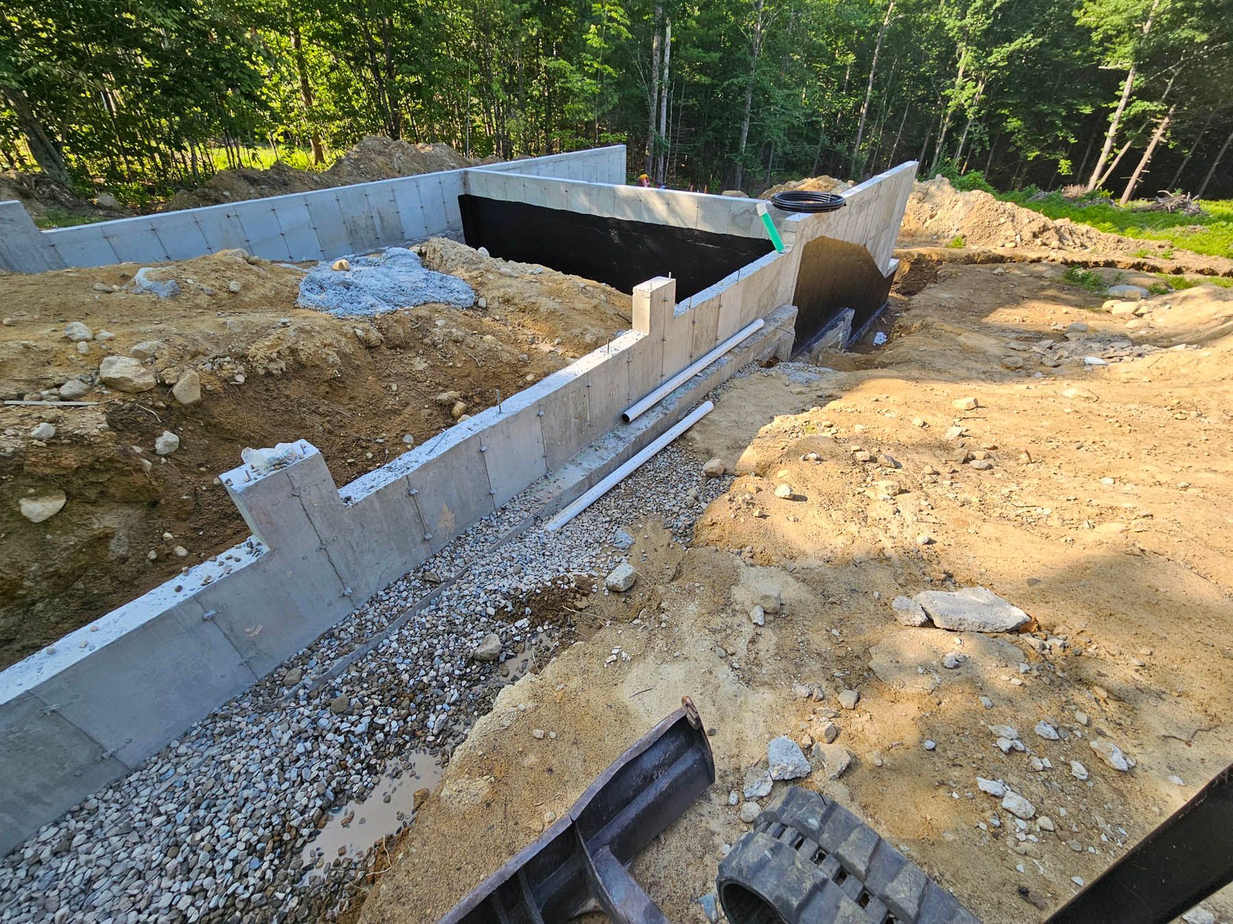 Foundation of a building under construction. Concrete walls, gravel base, black membrane, and surrounding dirt.