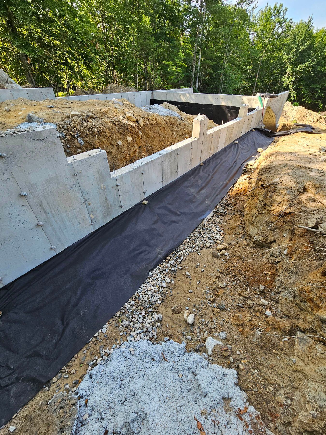 Concrete foundation with black fabric and gravel, partially buried in earth, near trees.