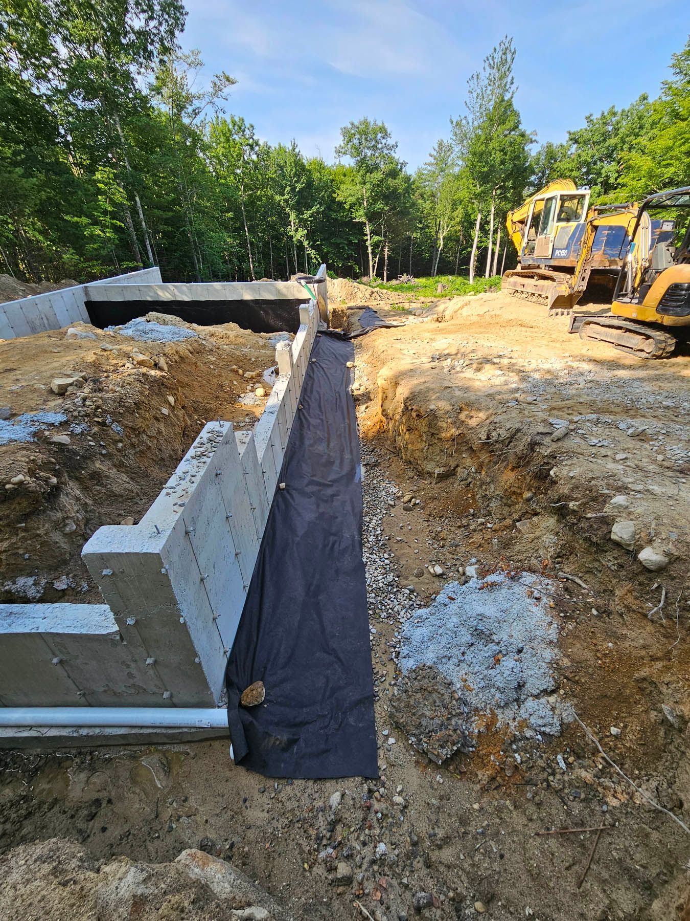 Construction site with a concrete foundation wall, earth, and heavy machinery; trees in the background.