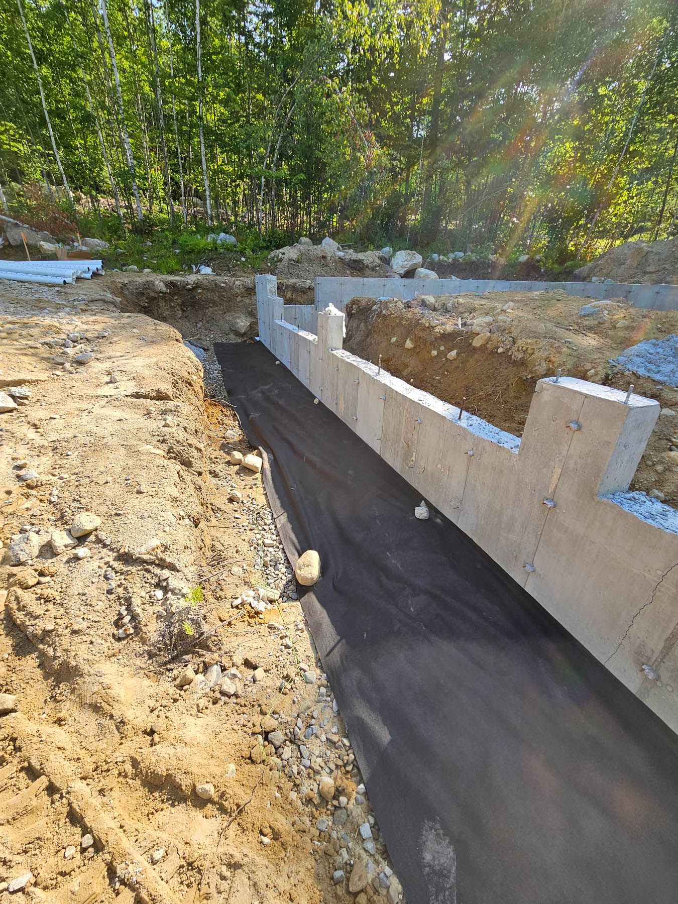 Construction site with concrete foundation wall and black fabric lining a trench, trees in the background.