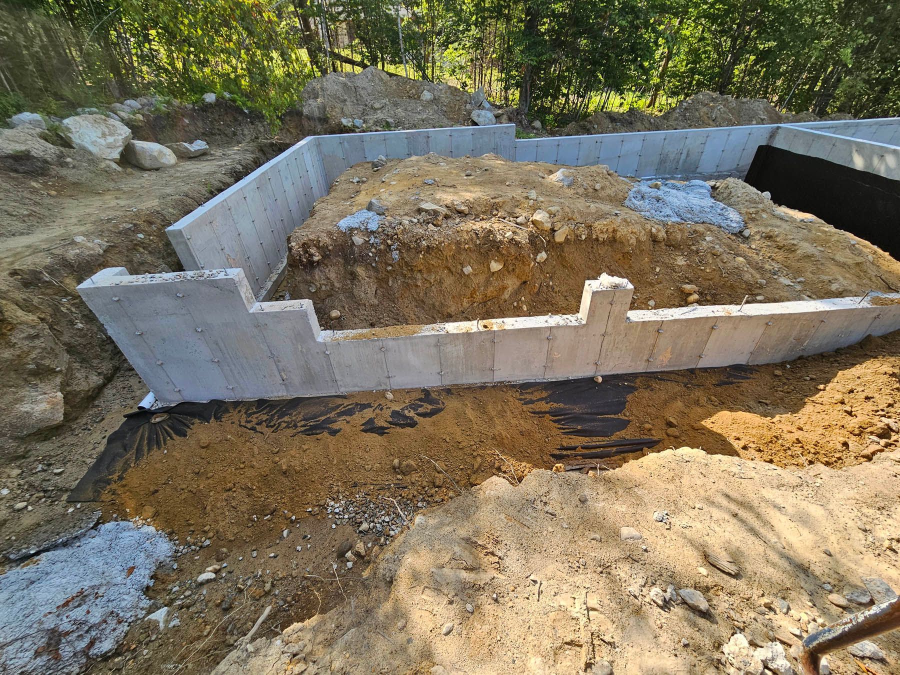 Concrete foundation walls, partially backfilled with soil, on a construction site.