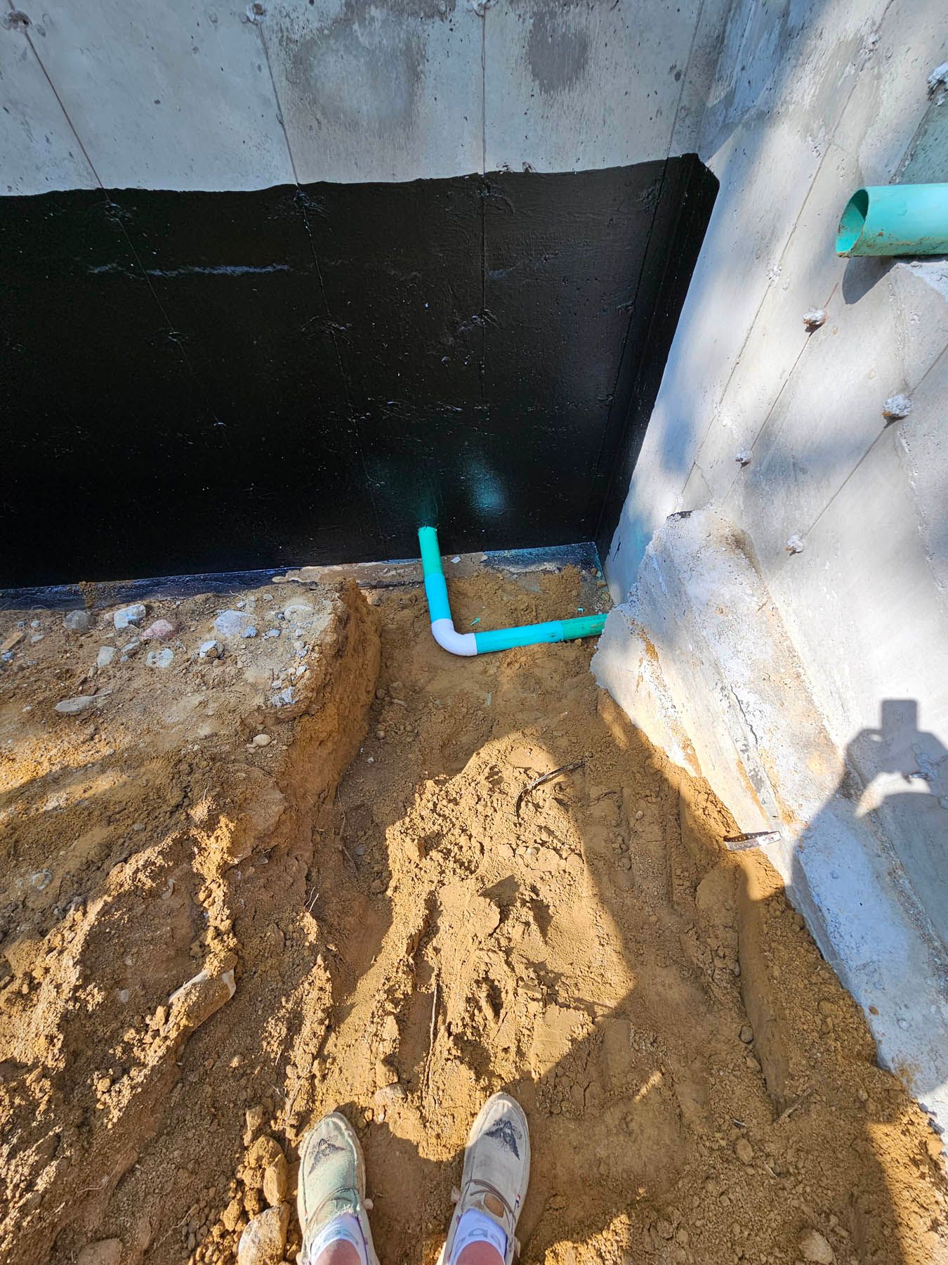 A person's feet, trench, and green pipes against a concrete wall with black waterproofing.