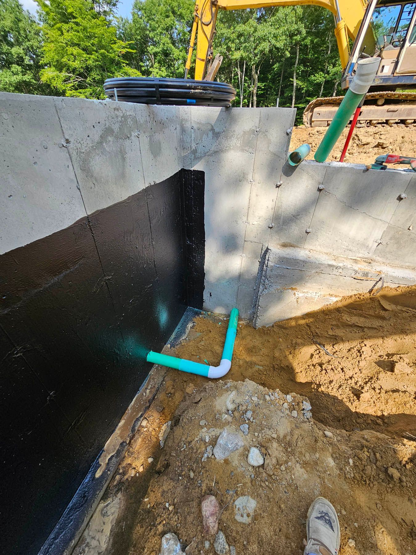 Foundation with black waterproofing, green pipes, and dirt. Construction site, sunny day.