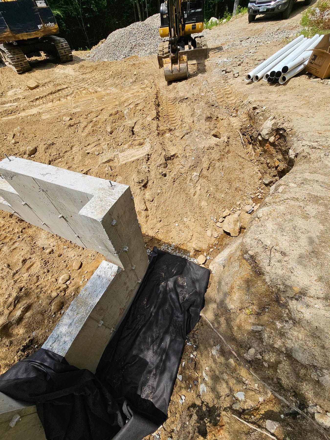 Construction site: excavator digging near concrete foundation, soil, and pipes.