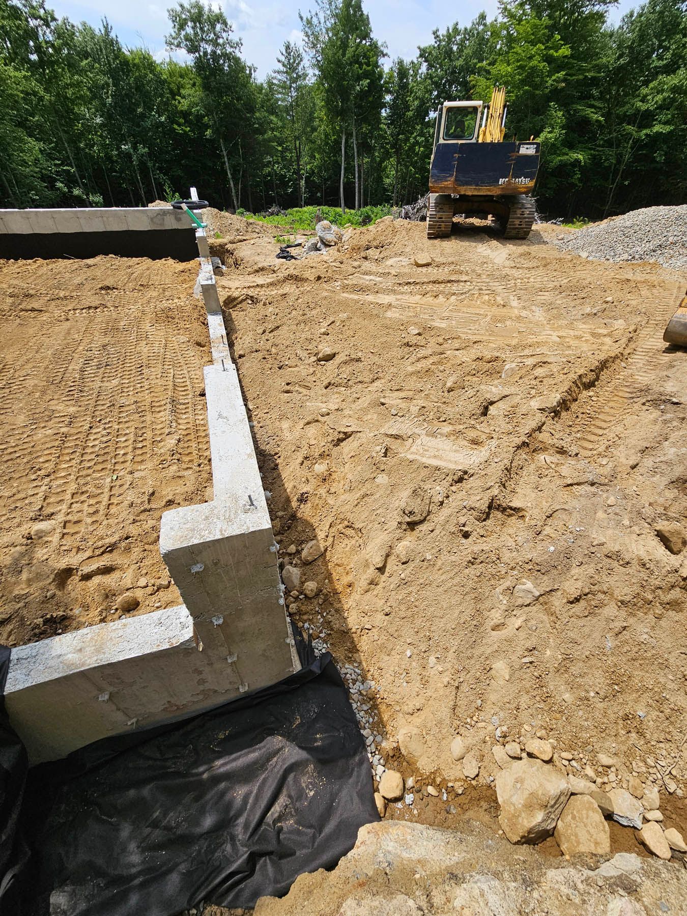 Construction site: concrete foundation walls with backhoe on dirt ground in a wooded area.
