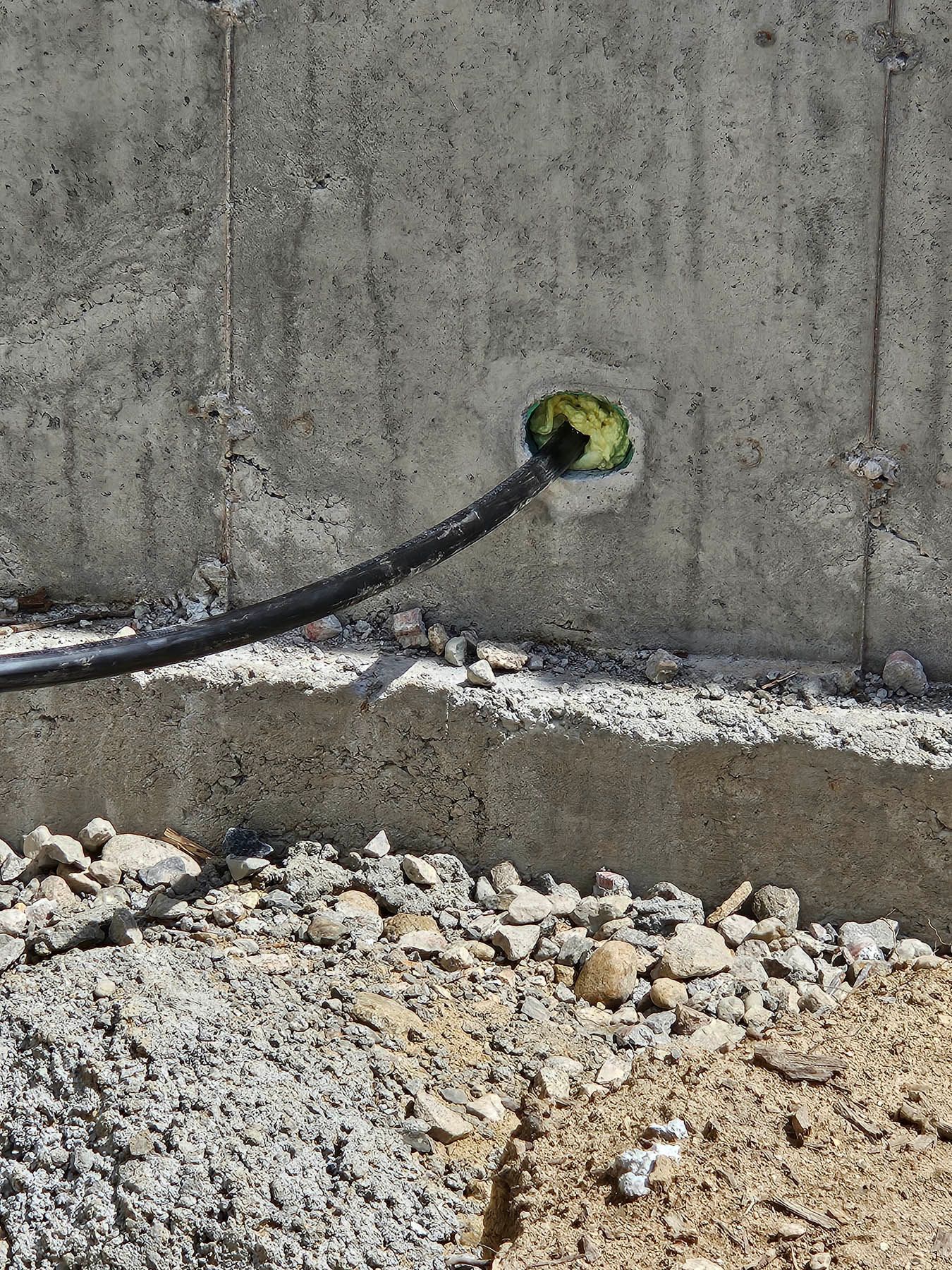 Black cable emerging from a hole in a concrete wall. Yellow sealant visible. Ground with gravel.