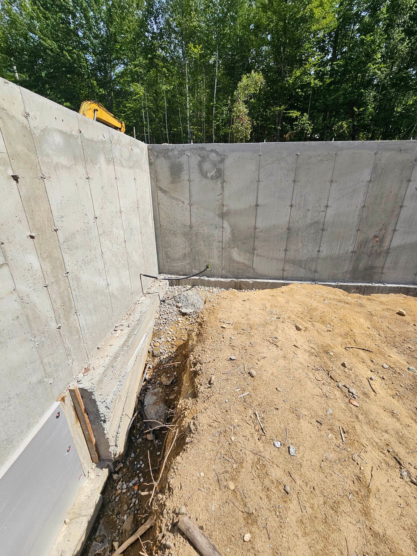 Concrete foundation corner next to dirt and trench. Yellow excavator in the background. Green trees.