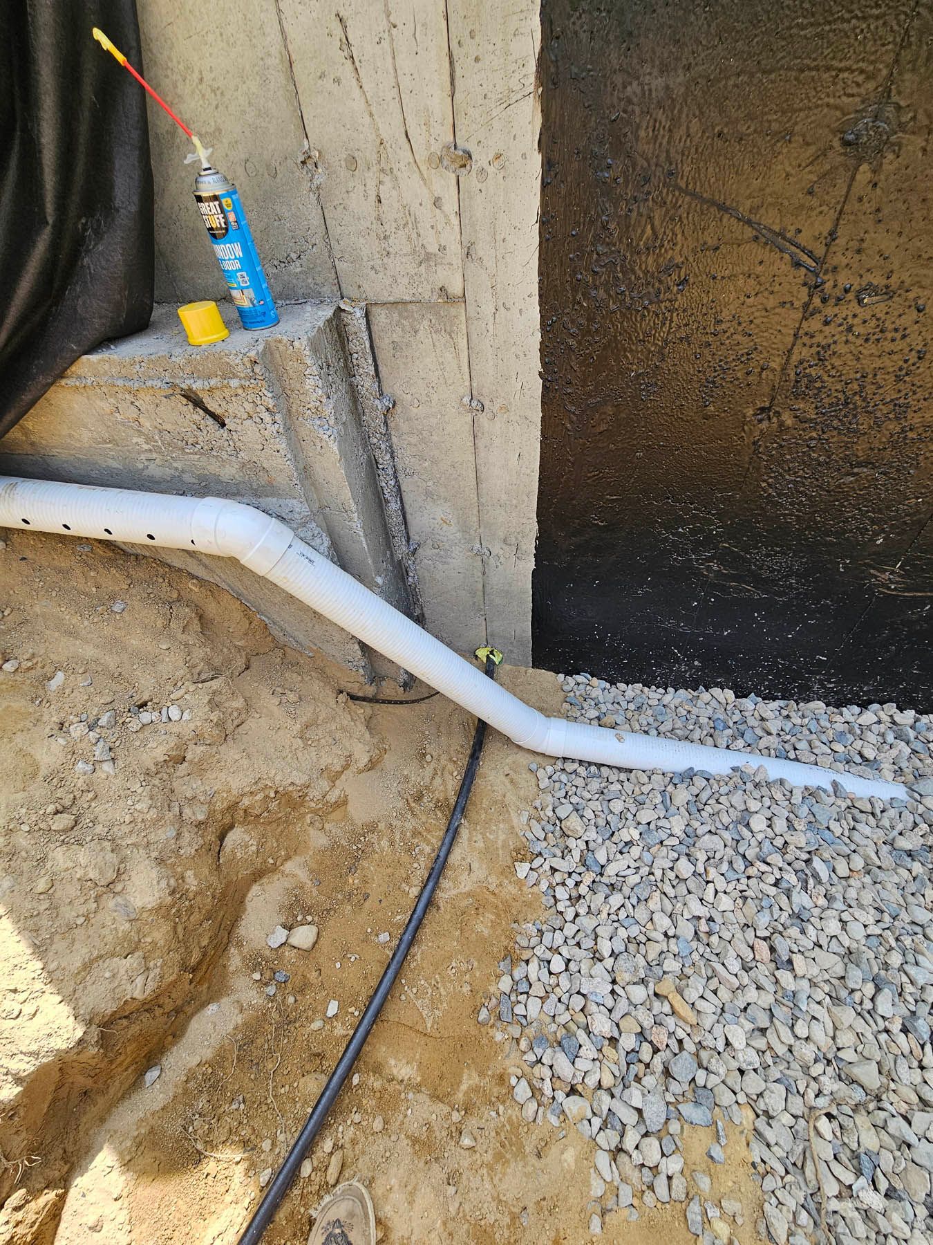 Foundation drainage system being installed with gravel and black waterproofing on wall.
