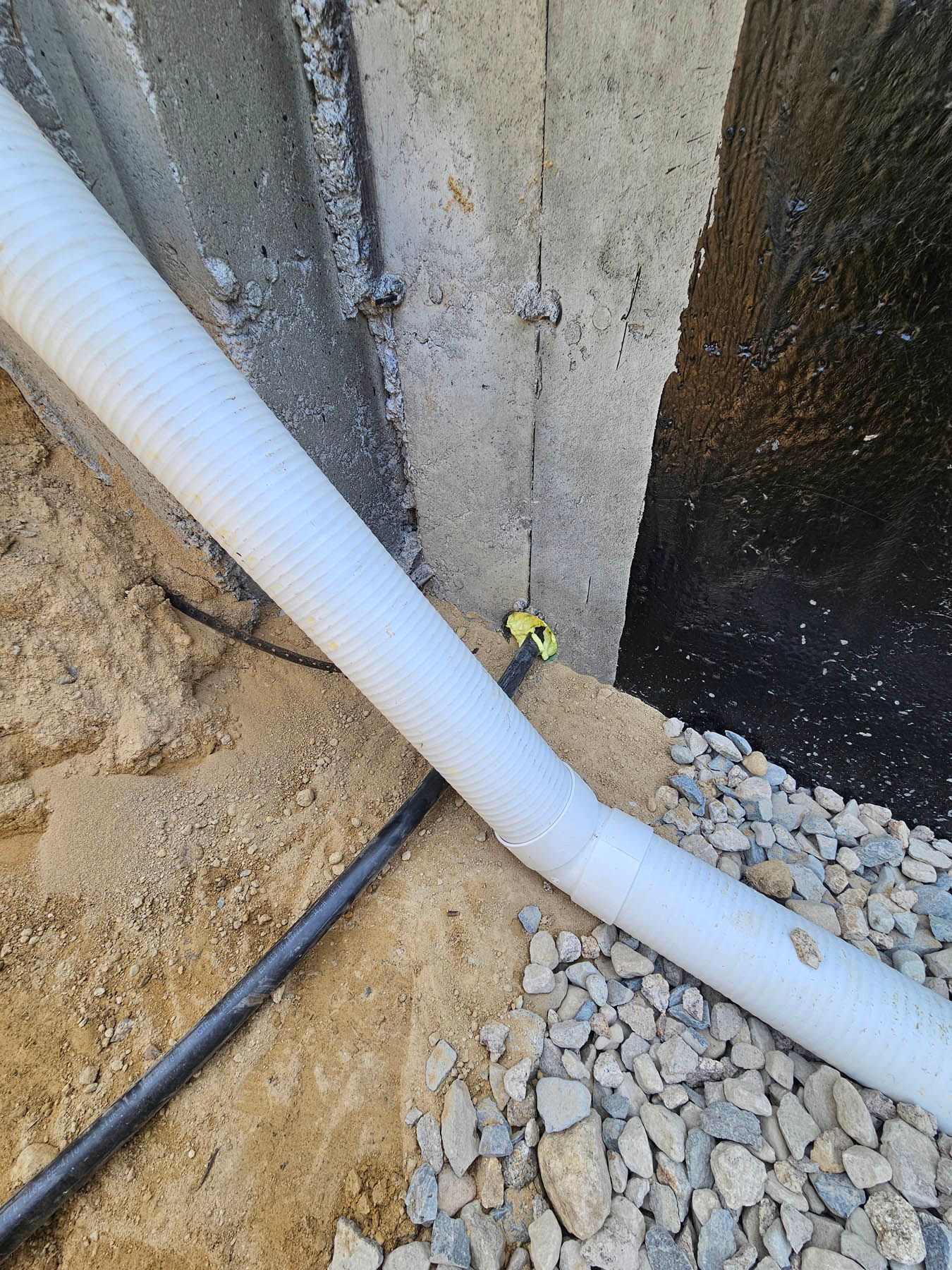 White corrugated pipe and black hose against a concrete foundation with gravel and sand.